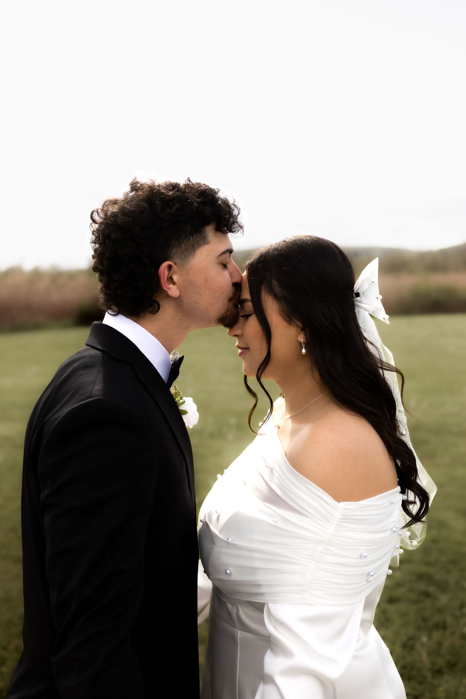 Groom gently kissing bride on the forehead as she smiles in a white off-shoulder wedding dress.