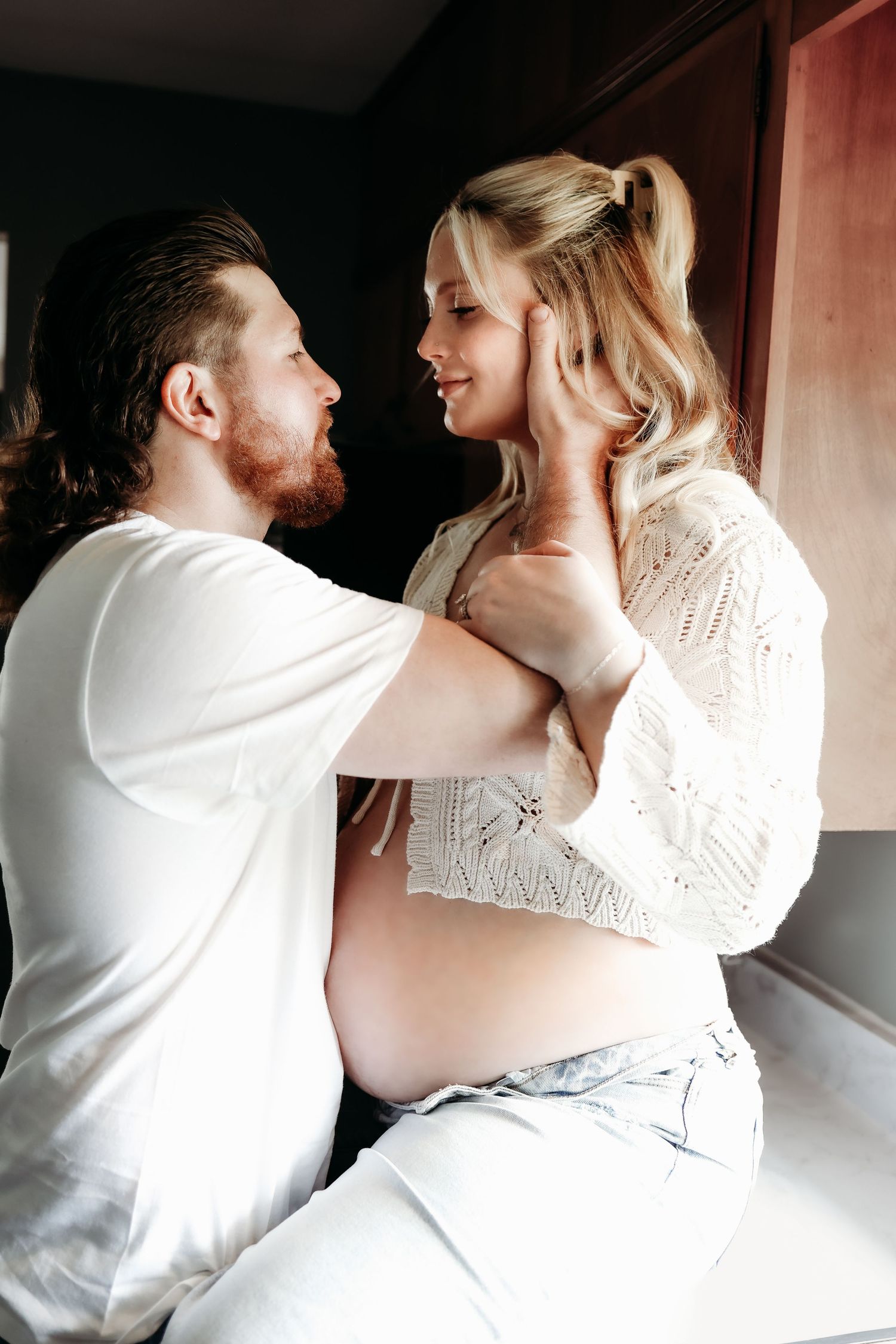 Tender moment between a bearded man and pregnant blonde woman in white outfits sharing an intimate look.