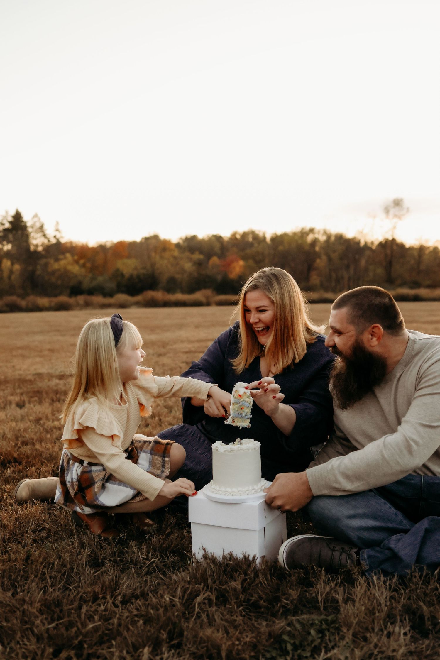 Family celebrating with a white birthday cake in an autumn field at golden hour sunset.