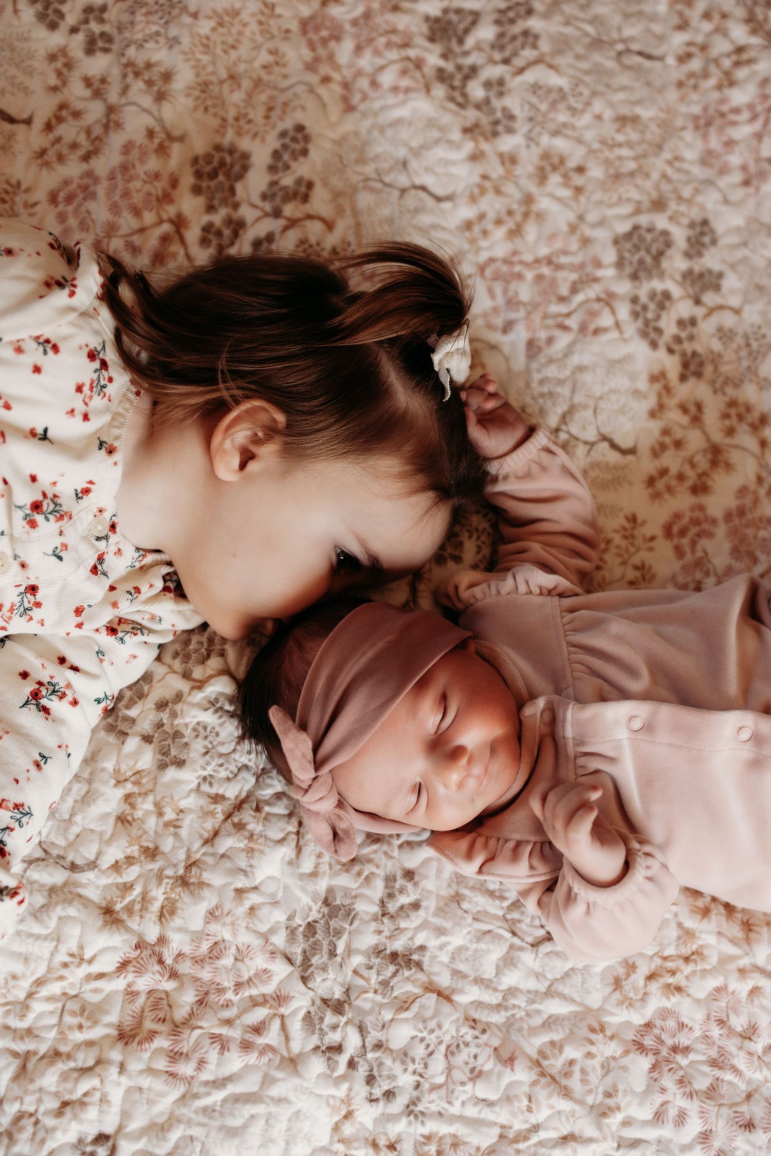 Young girl lying face-to-face with sleeping newborn sibling in pink outfit on floral quilt.
