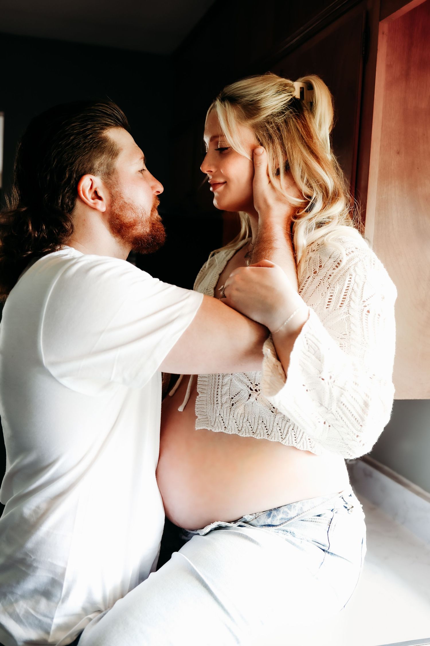 An intimate maternity portrait captures a tender moment between expectant parents in warm lighting.