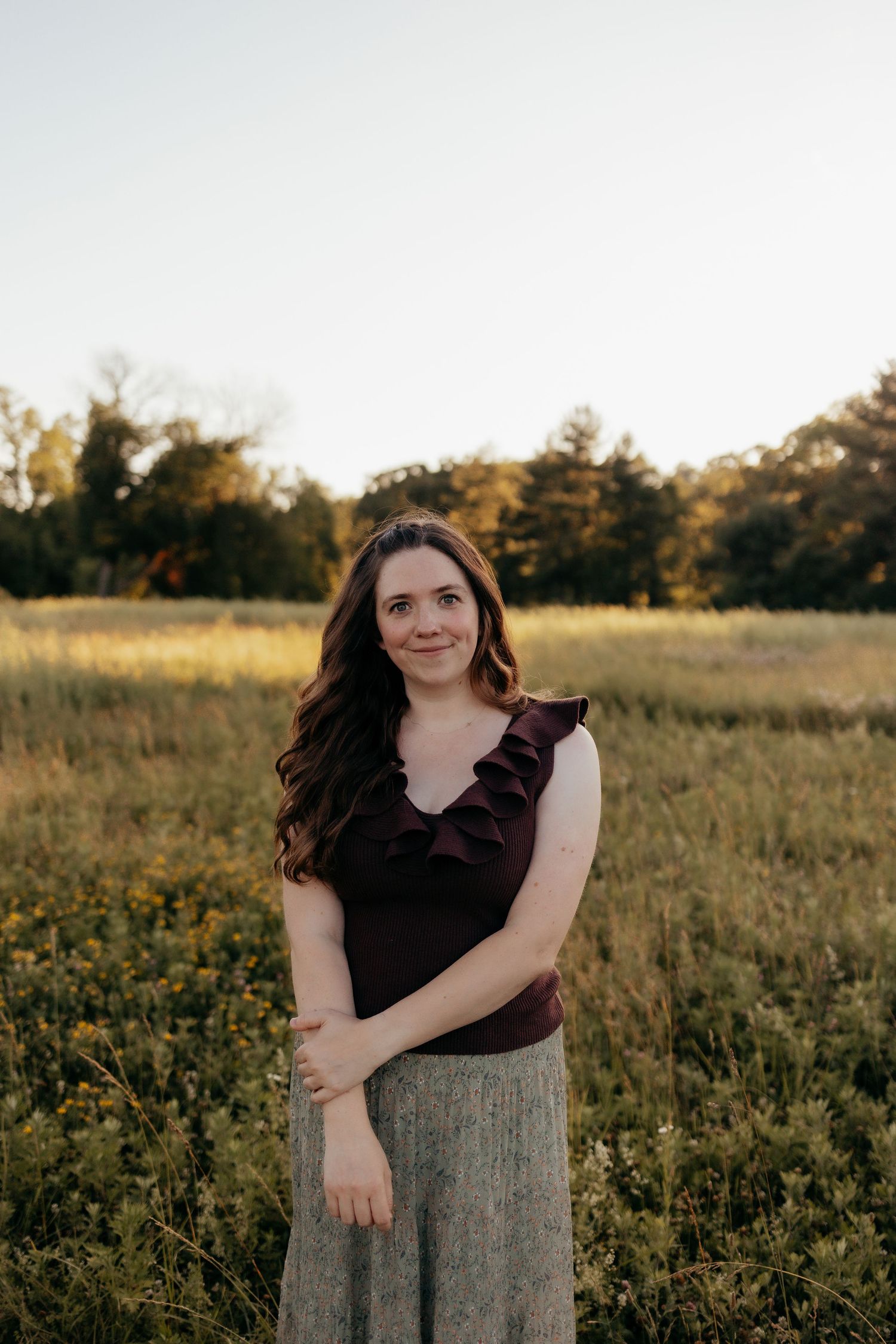 Young woman in burgundy ruffled top and maxi skirt smiles softly in a wildflower meadow at golden hour.