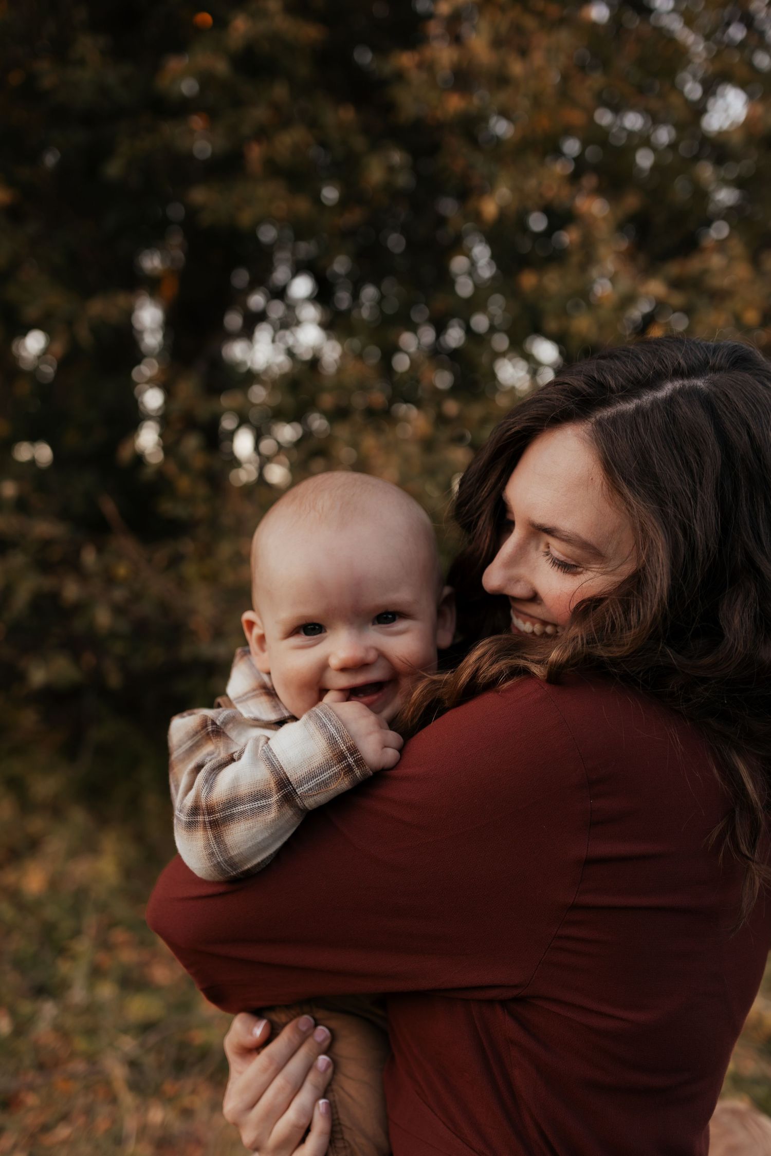 Smiling woman in a maroon sweater holds a happy baby in a plaid shirt close to her in an autumn outdoor setting.