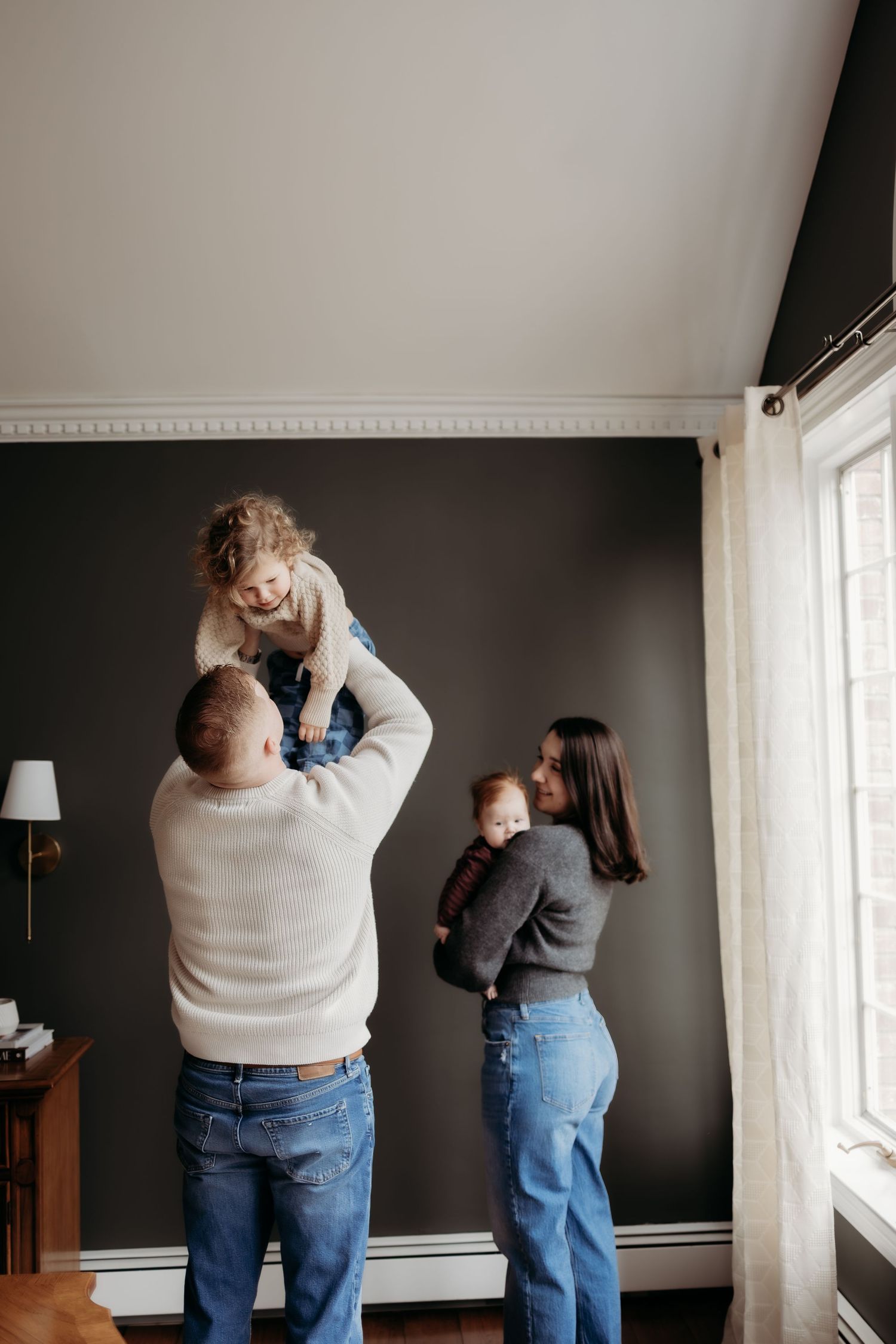 A happy family of four plays indoors, dad lifting toddler overhead while mom holds baby near a bright window.