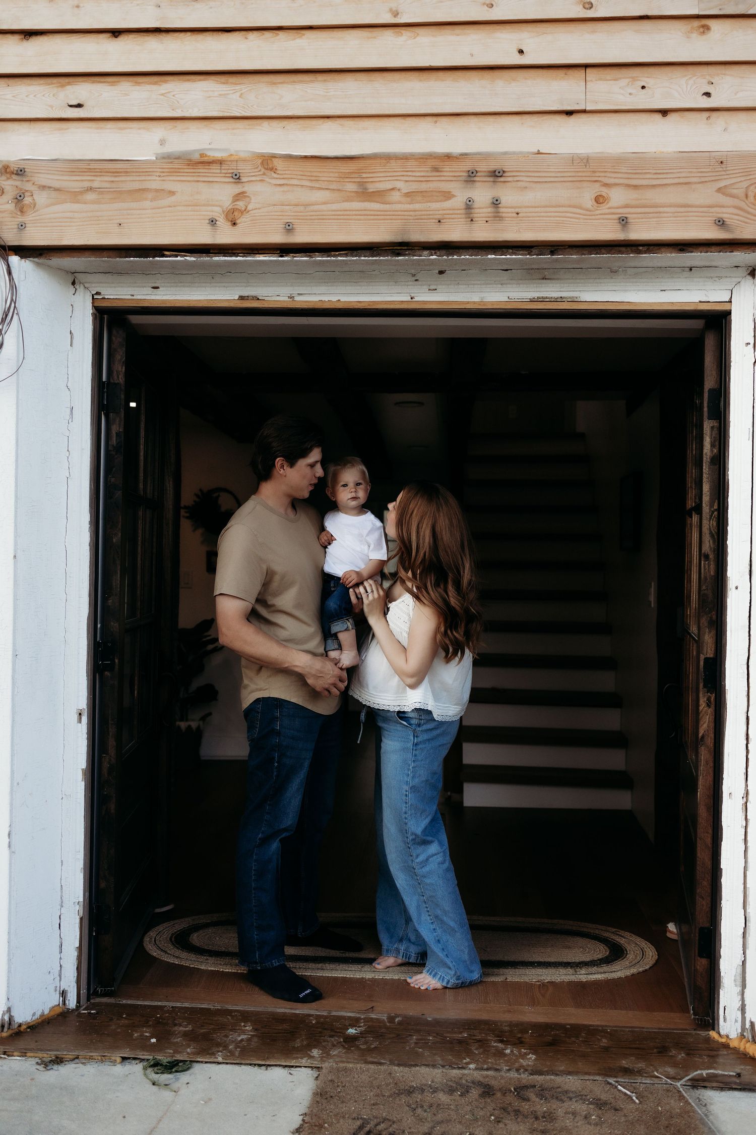 A young family with a toddler stands in an open garage doorway of a home under renovation.