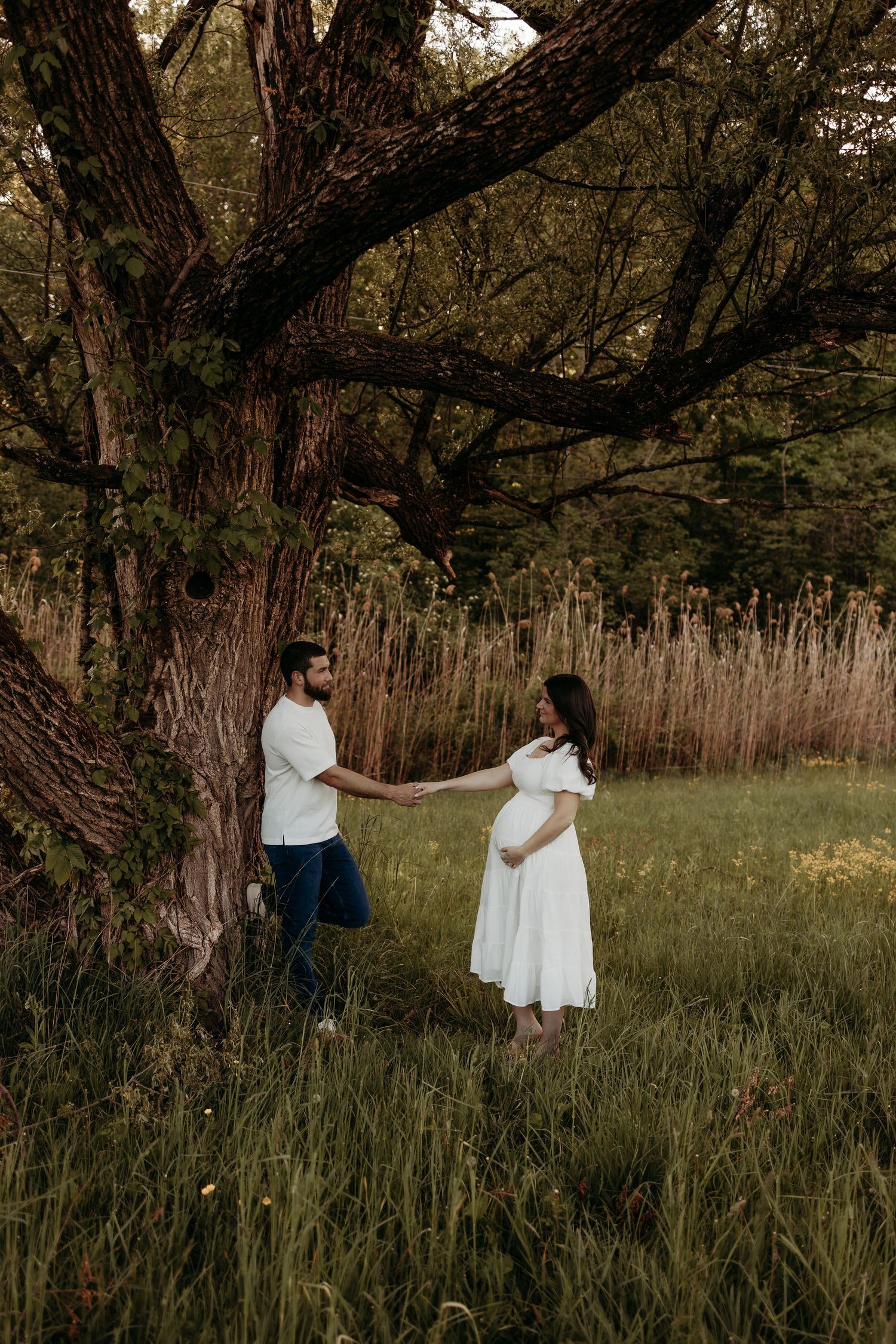Couple holding hands near large tree in lush green field at sunset.