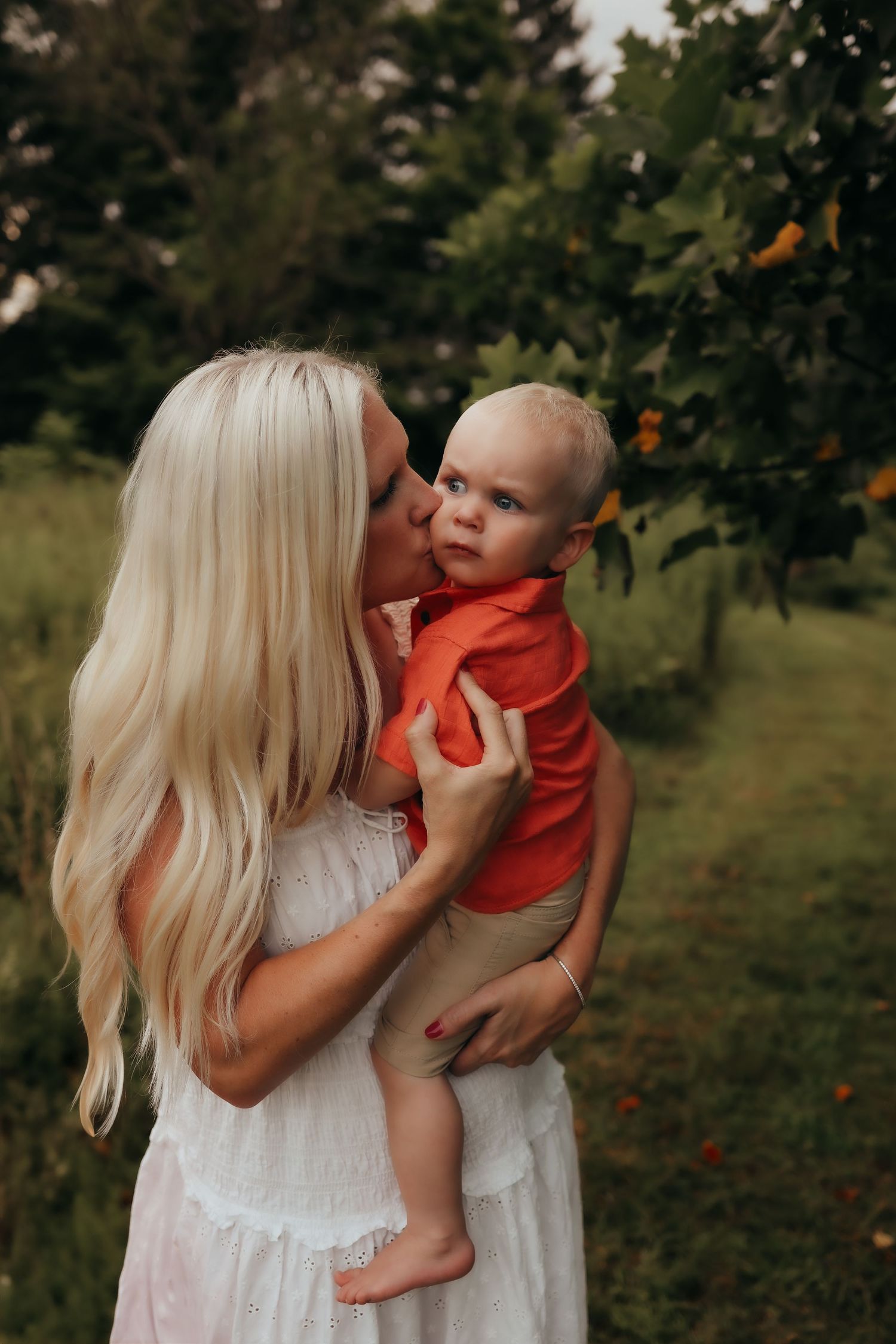 Blonde mother in white dress kissing baby boy in orange shirt outdoors near fruit trees.