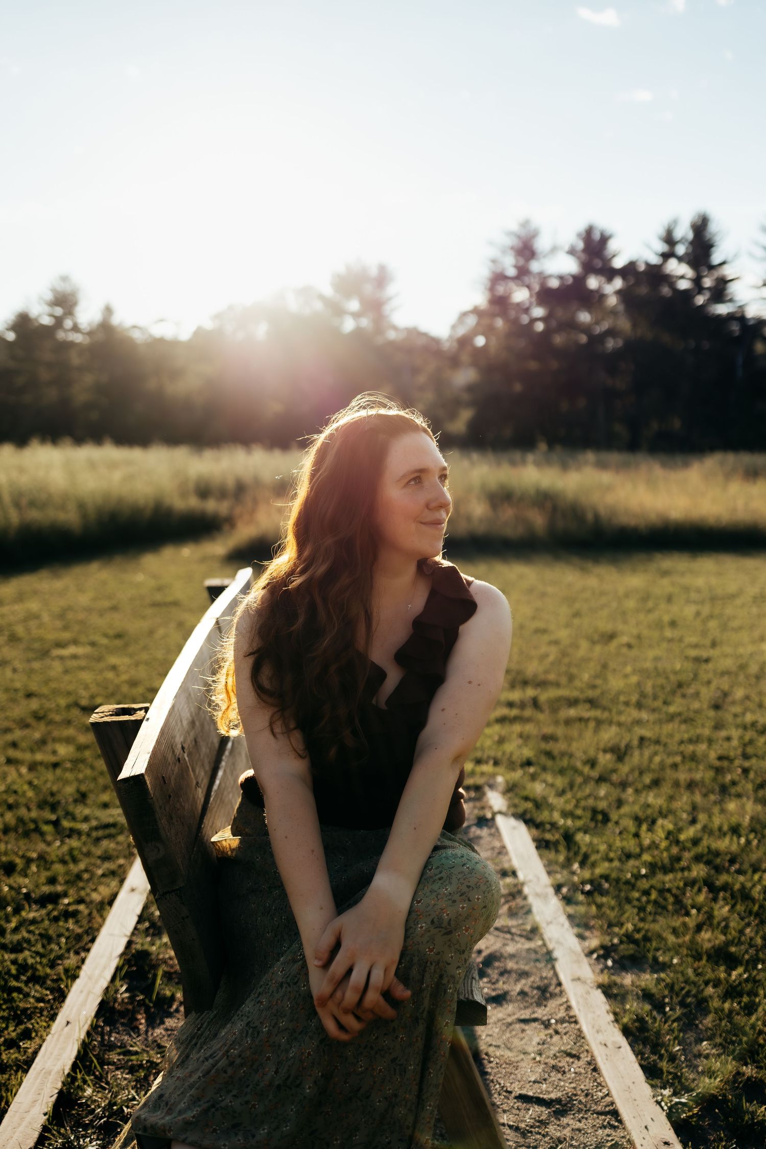Young woman with long auburn hair sits on wooden bench in sunlit field, smiling softly in golden light.