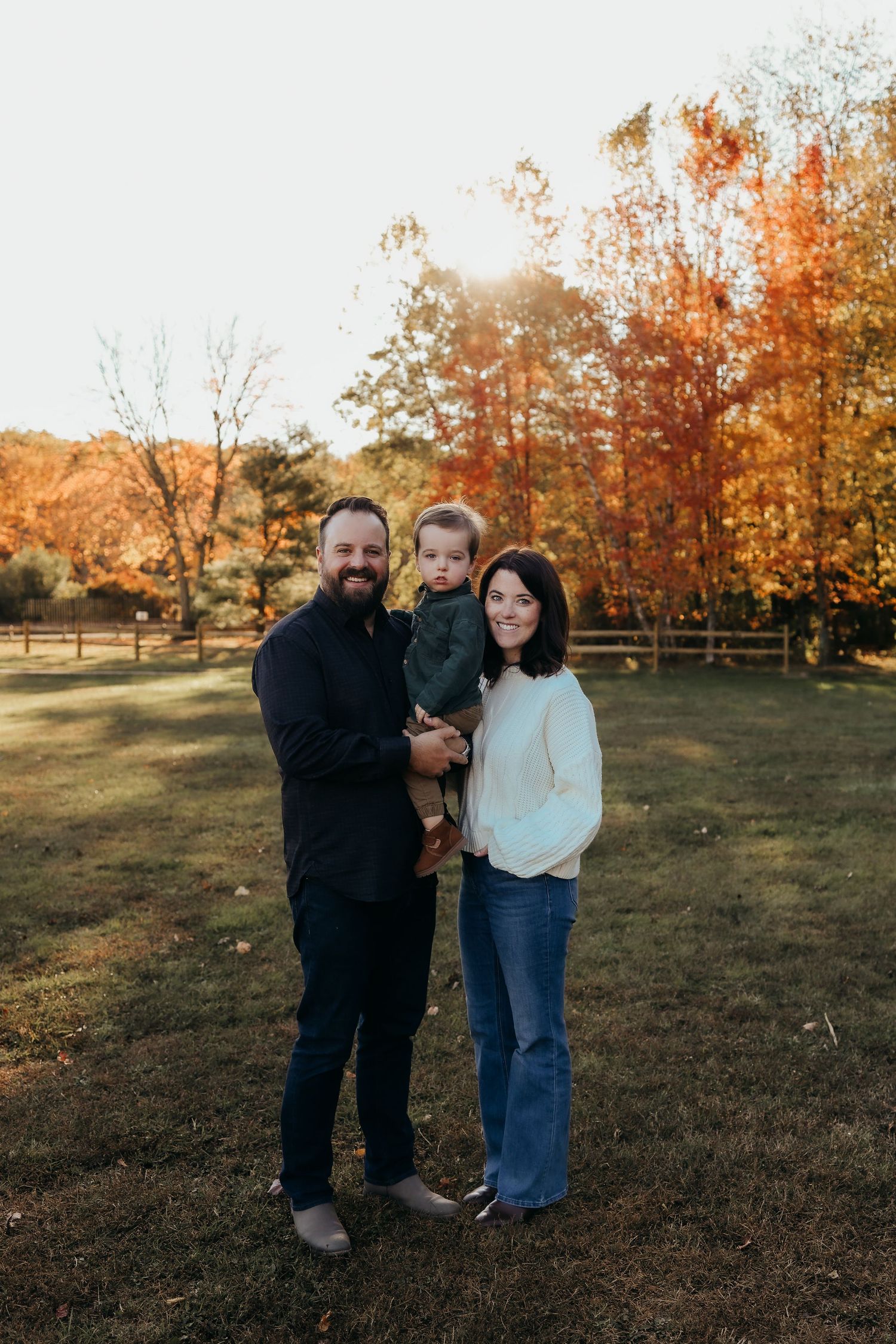 Family of three posing outdoors in autumn, parents holding toddler with colorful fall foliage and golden sunset in background.
