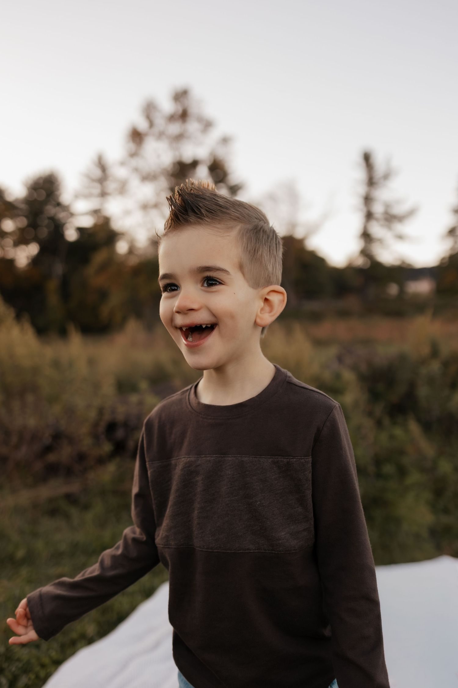 Smiling young boy with a spiky hairstyle laughs outdoors in a brown long-sleeve shirt, standing in a golden meadow at sunset.