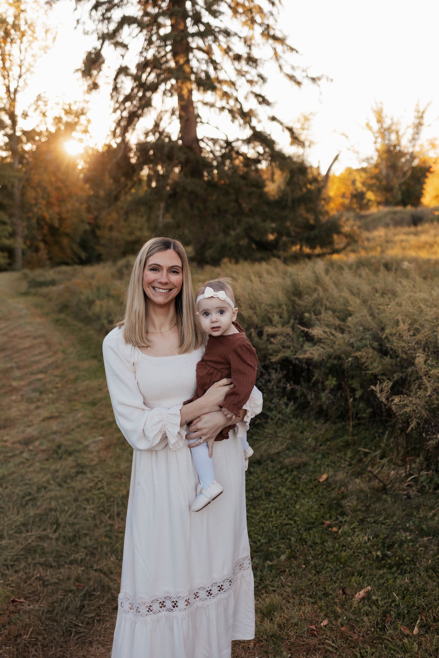 Smiling woman in a cream dress holds a baby in a rust outfit and bow headband in a golden-hour field with tall trees behind.