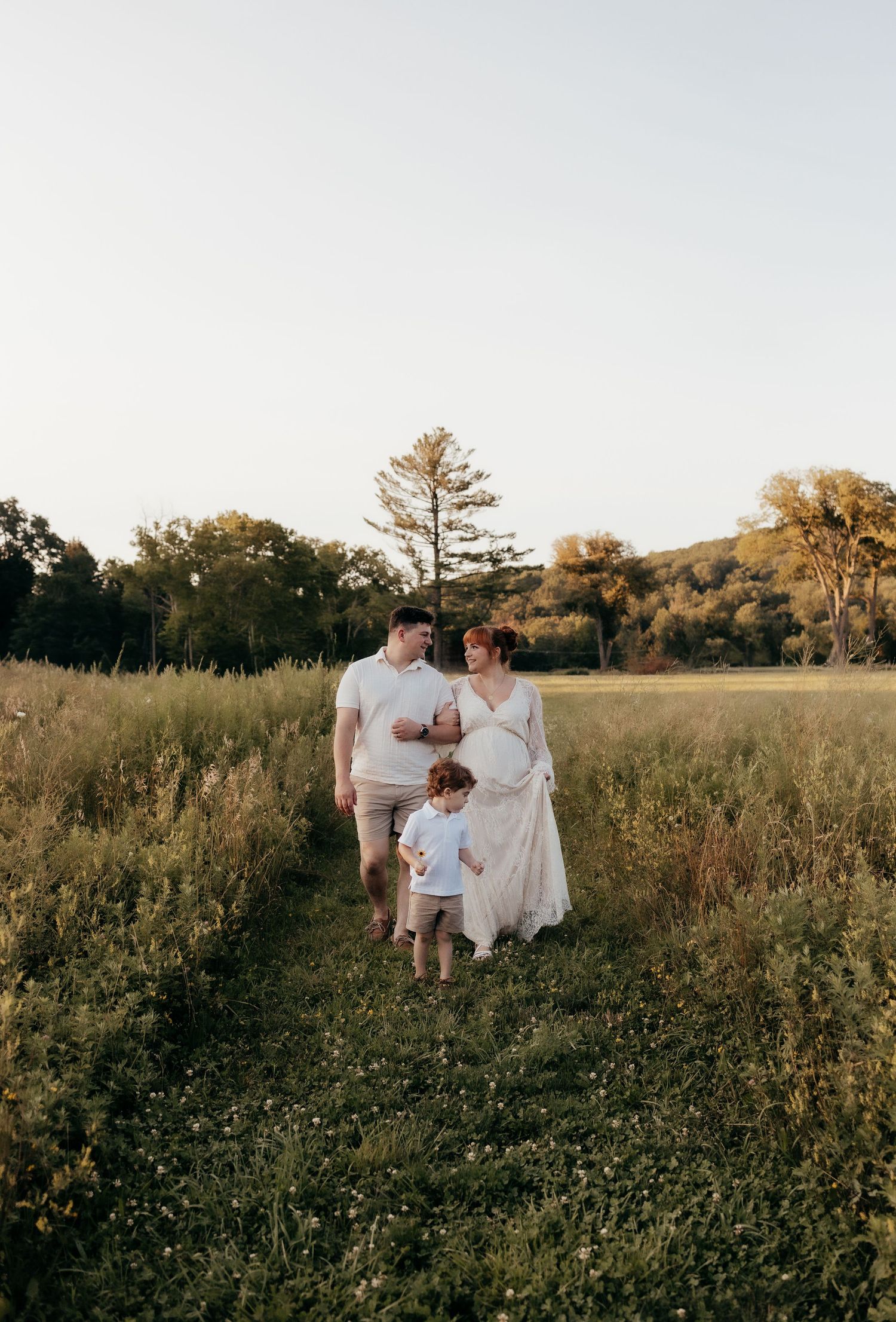 A family walks through a sunlit meadow path, with a pregnant woman in a white dress, a man, and a young child.