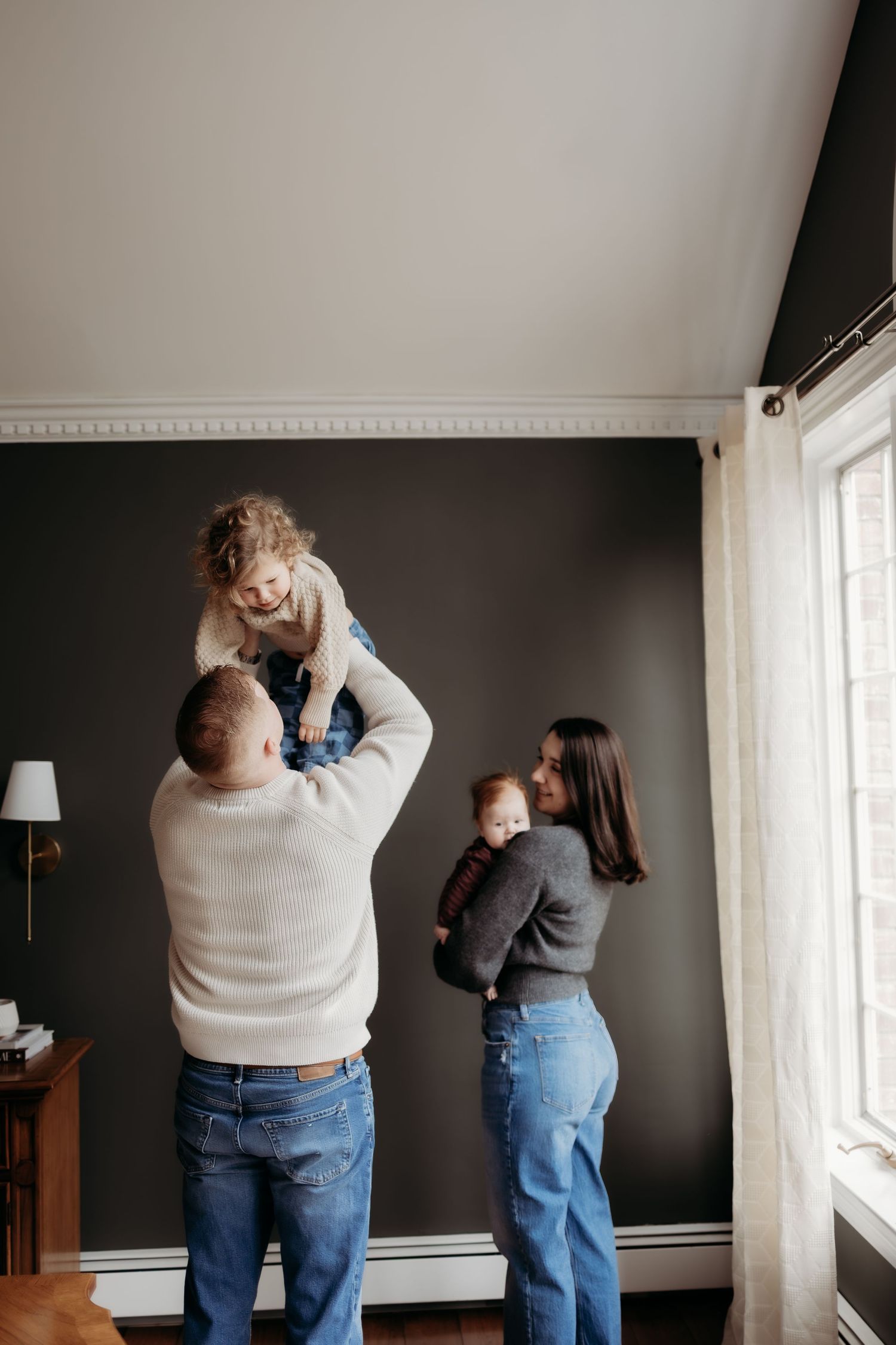 Parents play with two young children in a dark-walled living room, lifting toddler overhead.