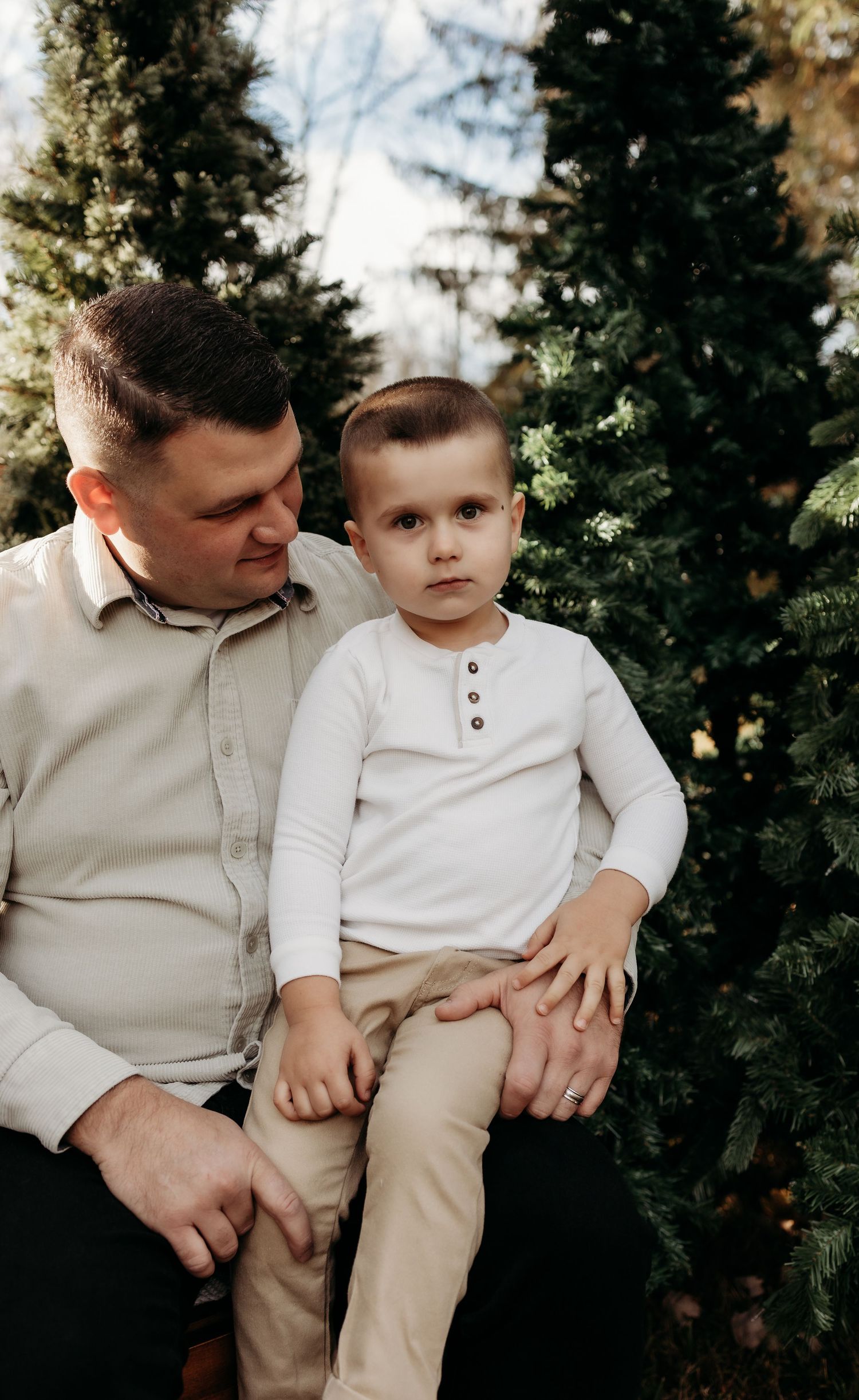 A father holds his young son on his lap outdoors surrounded by lush green evergreen trees in a warm family portrait.