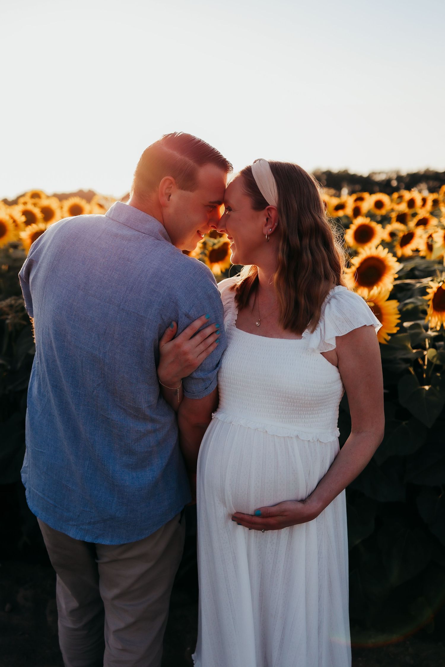 A couple shares romantic moment in a sunflower field, the woman wearing a white dress during a maternity photoshoot in Newton, New Jersey
