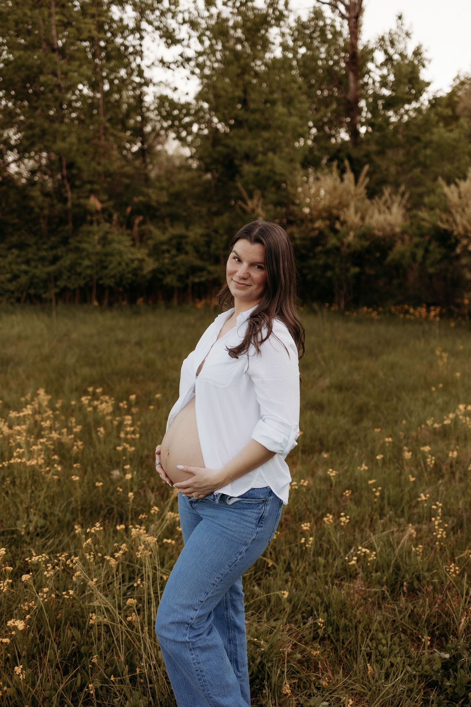 Smiling pregnant woman in white shirt and jeans cradling baby bump in a sunny wildflower meadow.