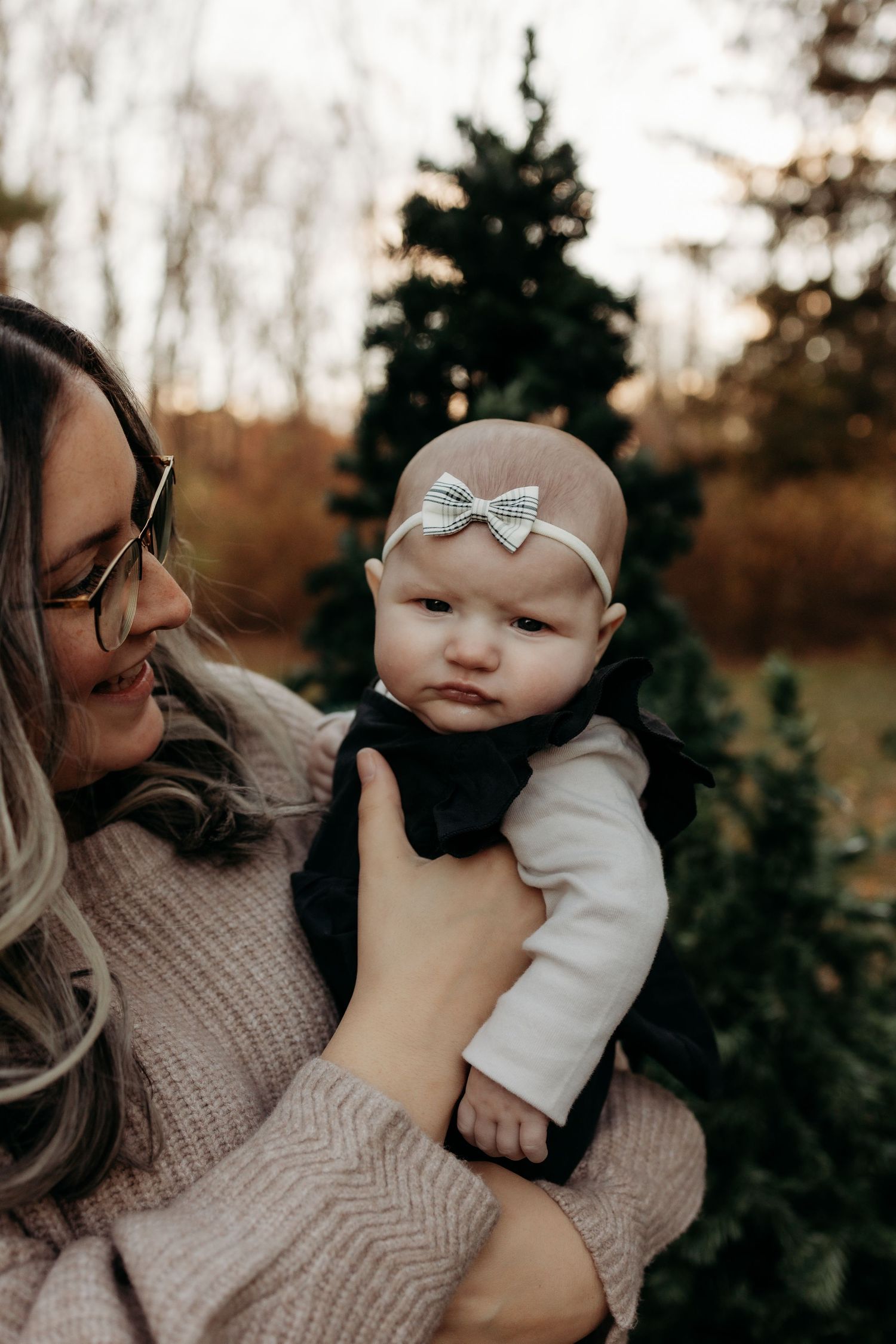 A mother in a pink sweater holds her baby girl wearing a bow headband outdoors near Christmas trees.