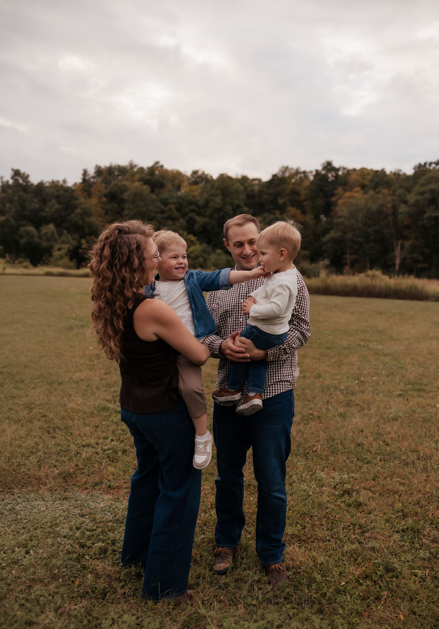 Family of four smiling outdoors in a green field, parents each holding a young child on a cloudy autumn day.