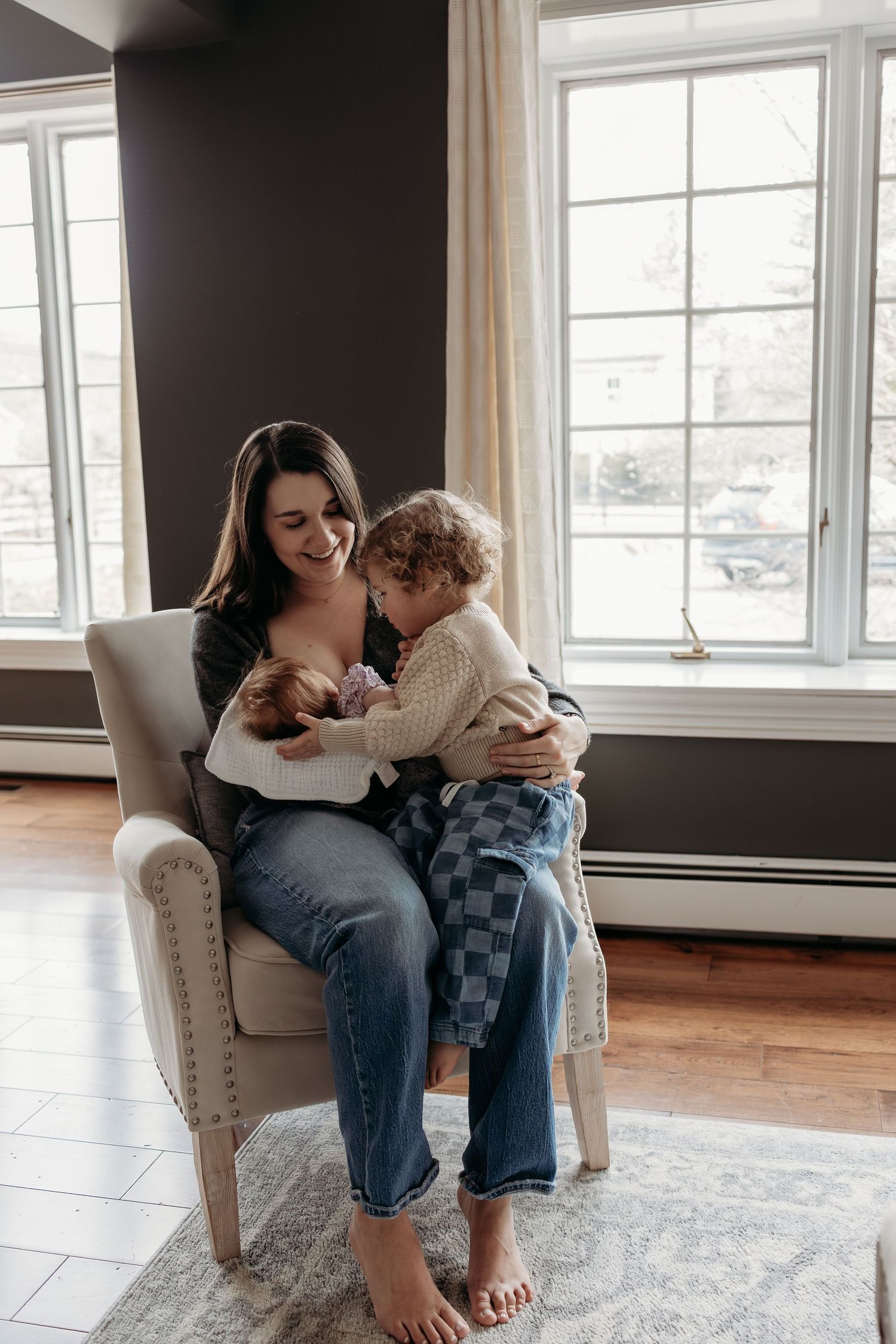 Smiling mother sitting in armchair breastfeeding newborn while toddler leans in for a cuddle by bright windows.