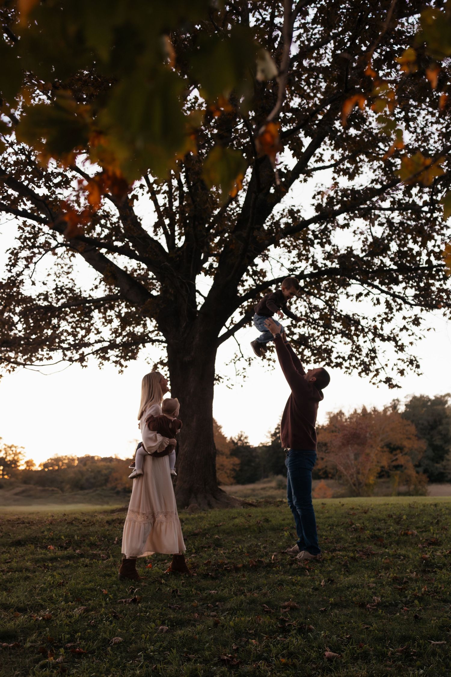 Family of three plays beneath a large autumn tree at sunset as a father lifts a toddler overhead while the mother watches.