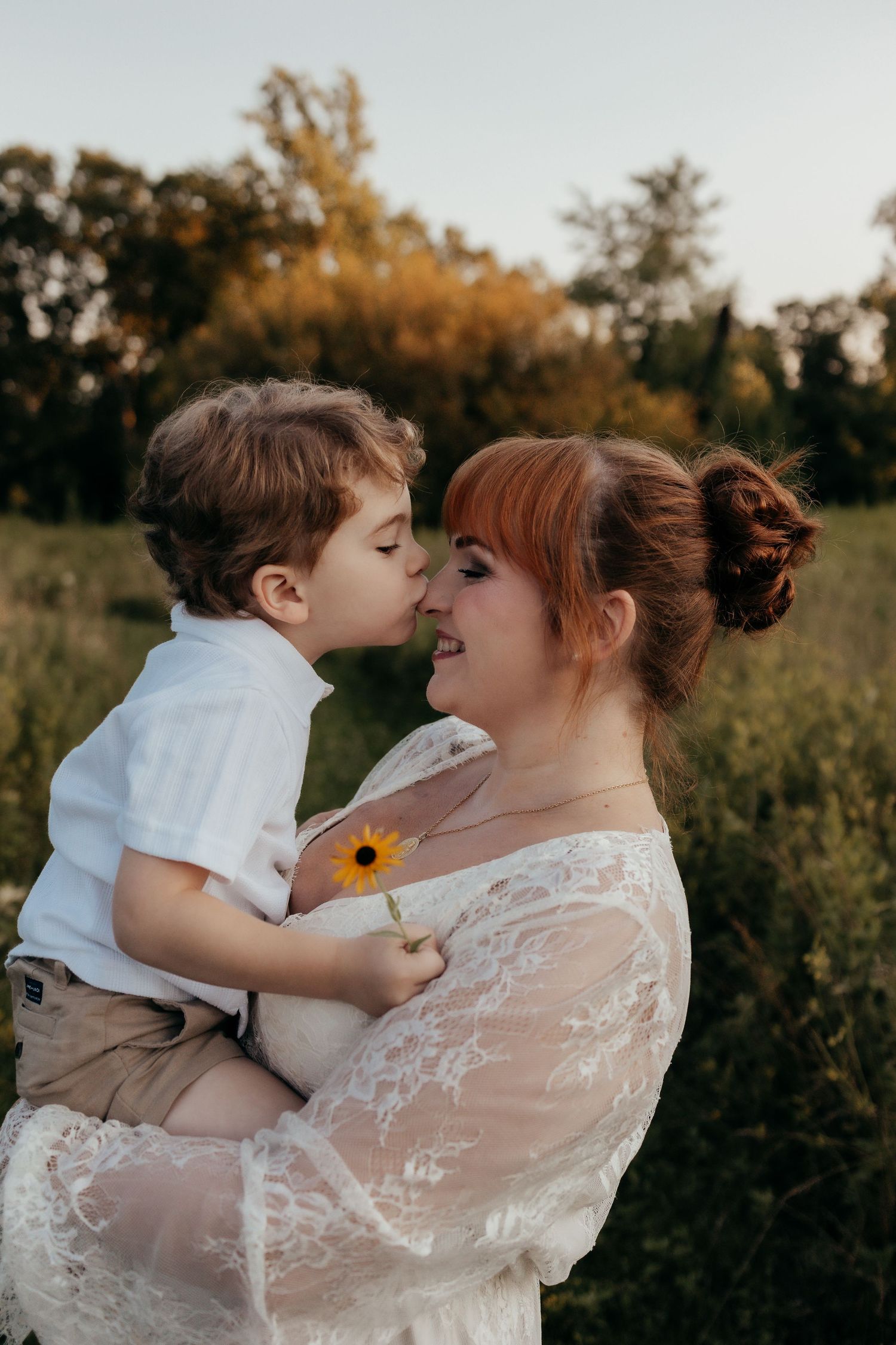 A young boy kisses his redheaded mother on the nose while holding a sunflower in a lush green field.
