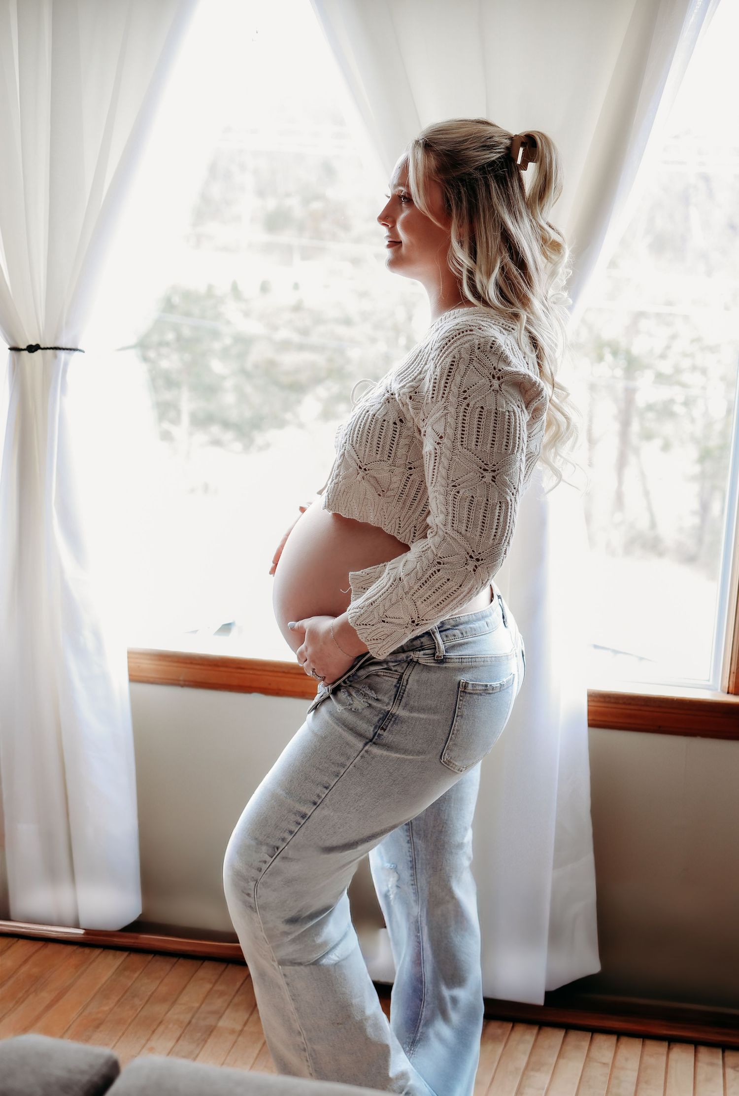 Side profile of pregnant blonde woman in knit top and jeans standing by window.