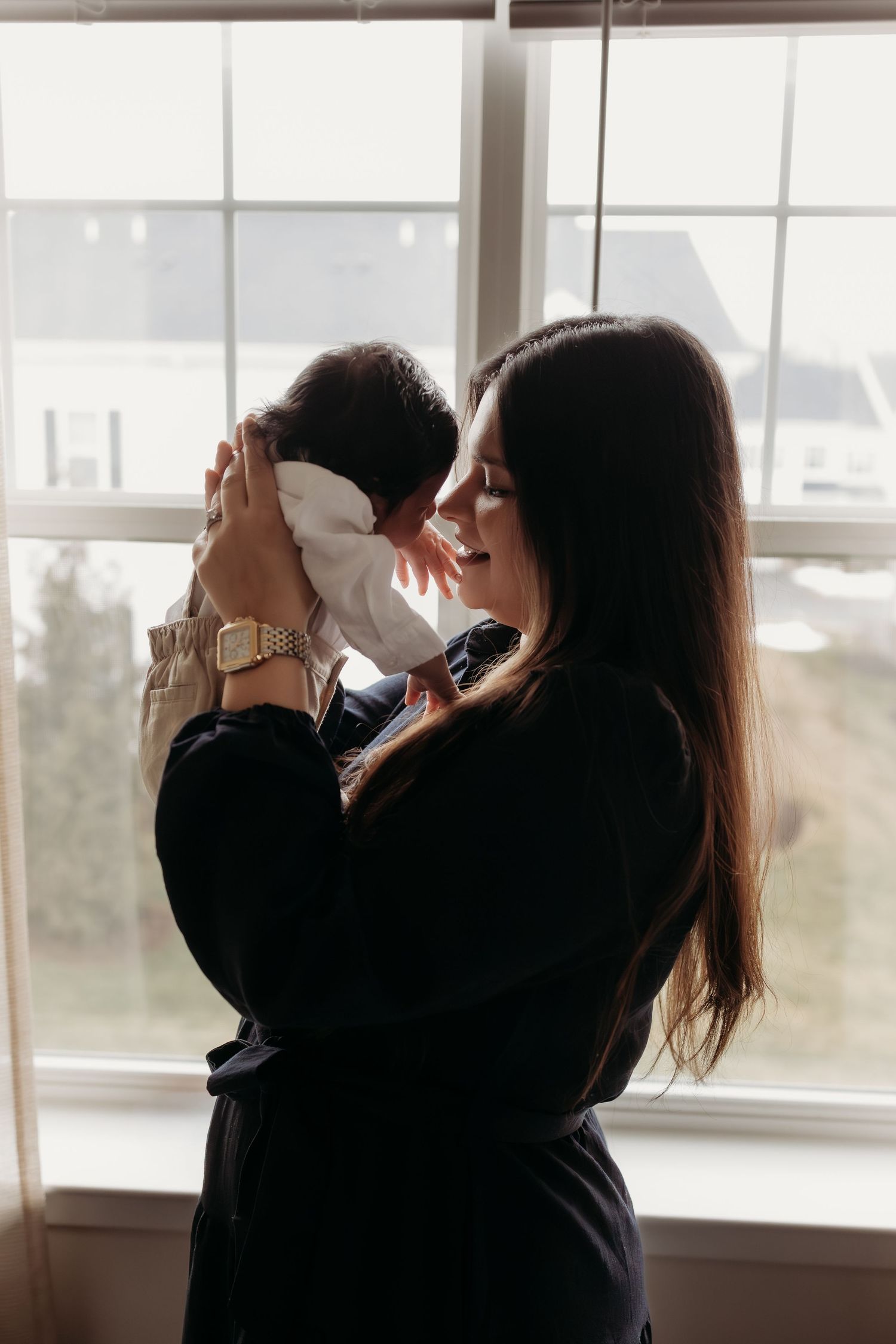 A mother lifts her newborn baby up to her face while standing in front of a bright window.