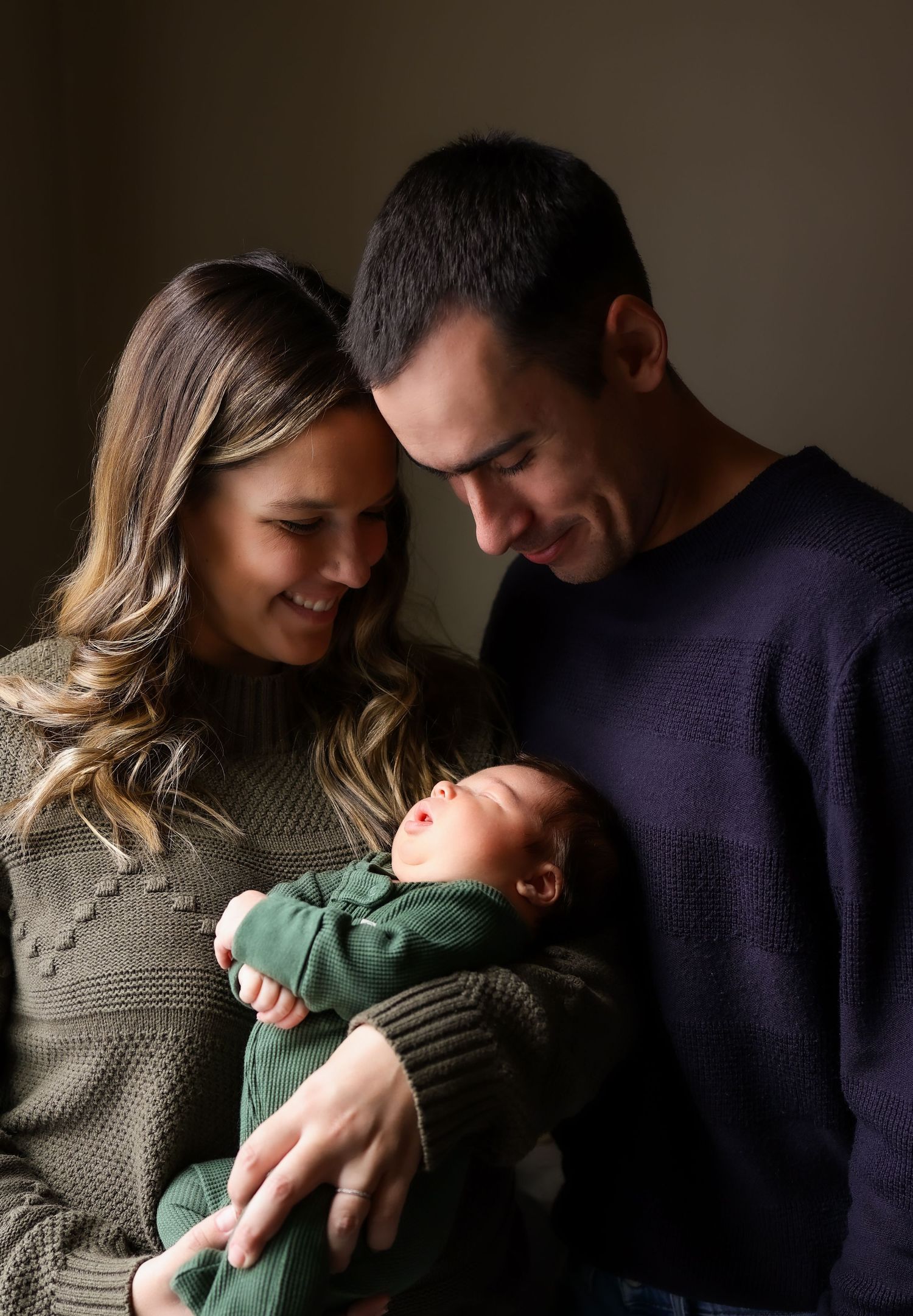 Loving parents smiling at newborn baby dressed in green outfit in moody portrait photo.