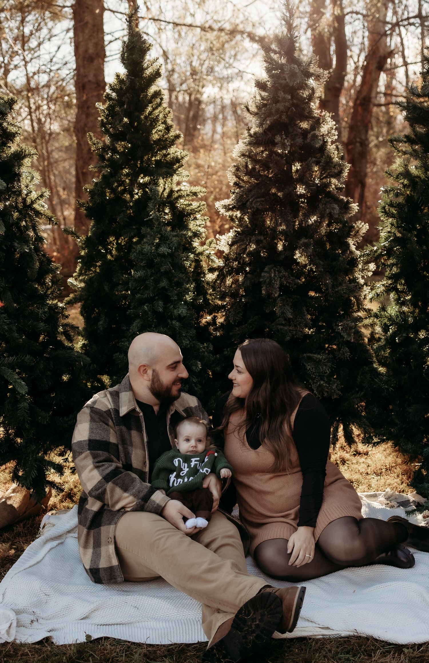 A family sits on a blanket among evergreen trees, with parents lovingly gazing at their baby in a holiday outdoor portrait.