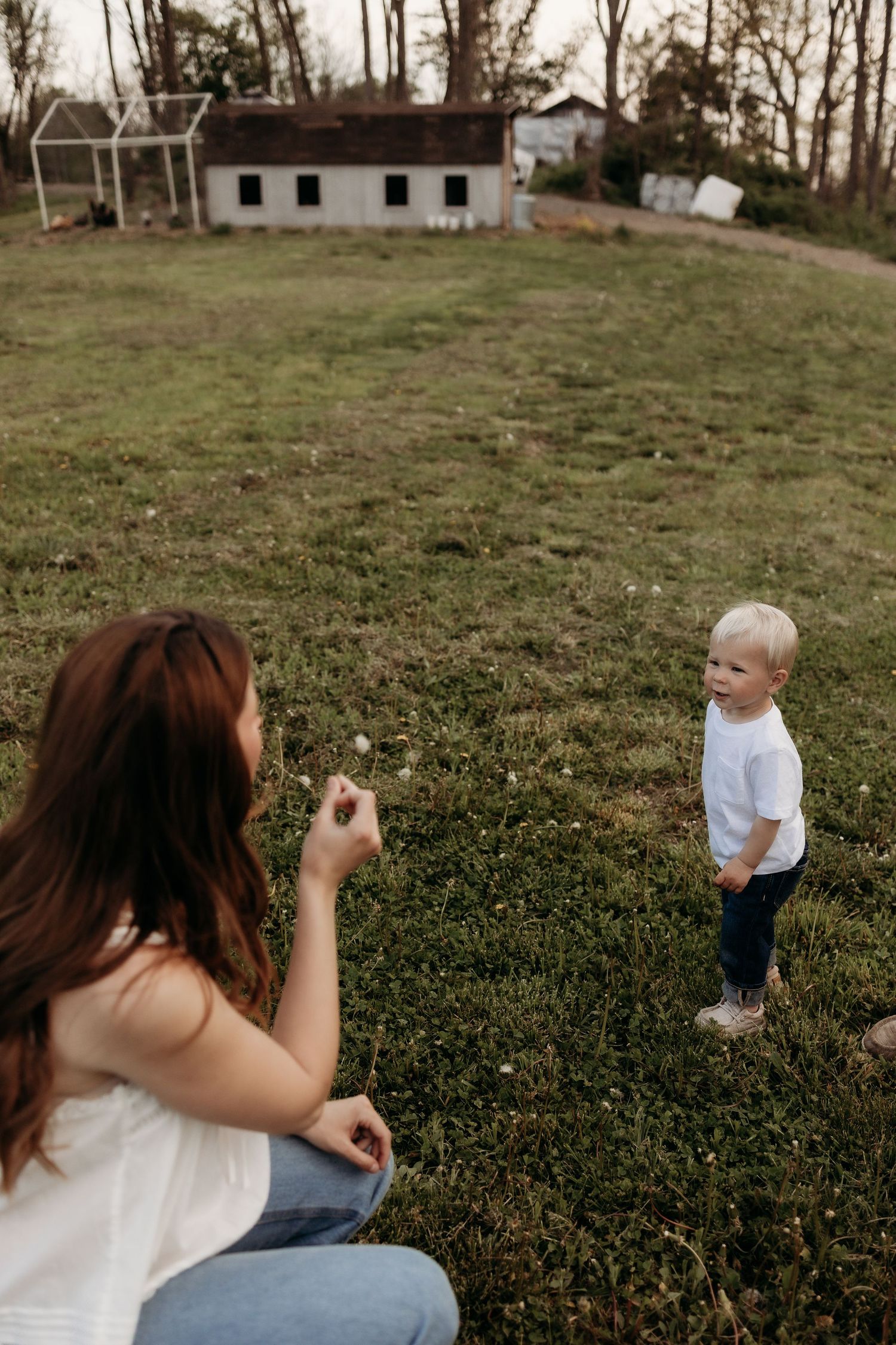 A mother crouches on a grassy field interacting with her young blonde toddler near a rustic farmhouse.
