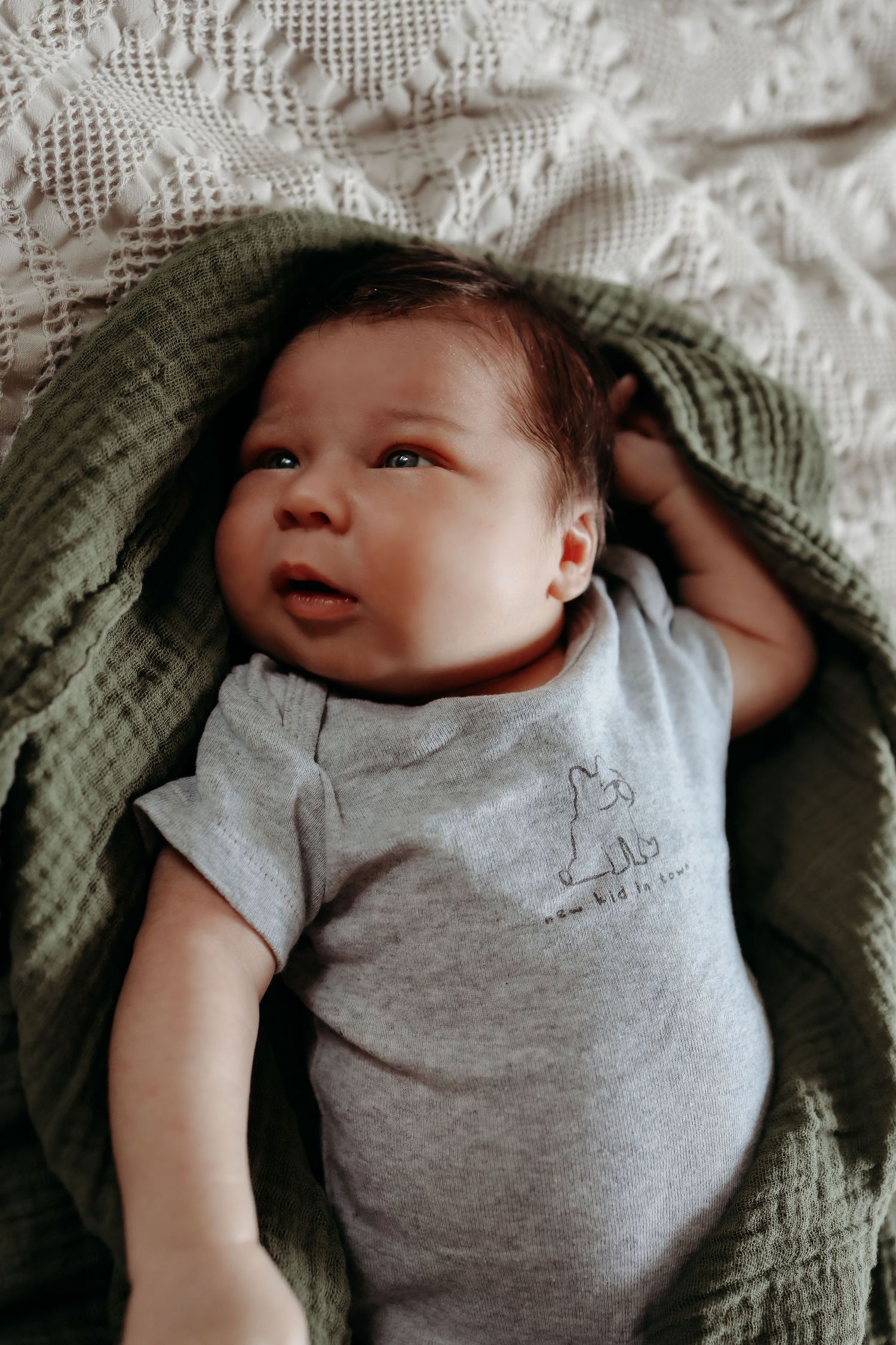 Adorable newborn baby in gray onesie lying on green muslin blanket looking upward.