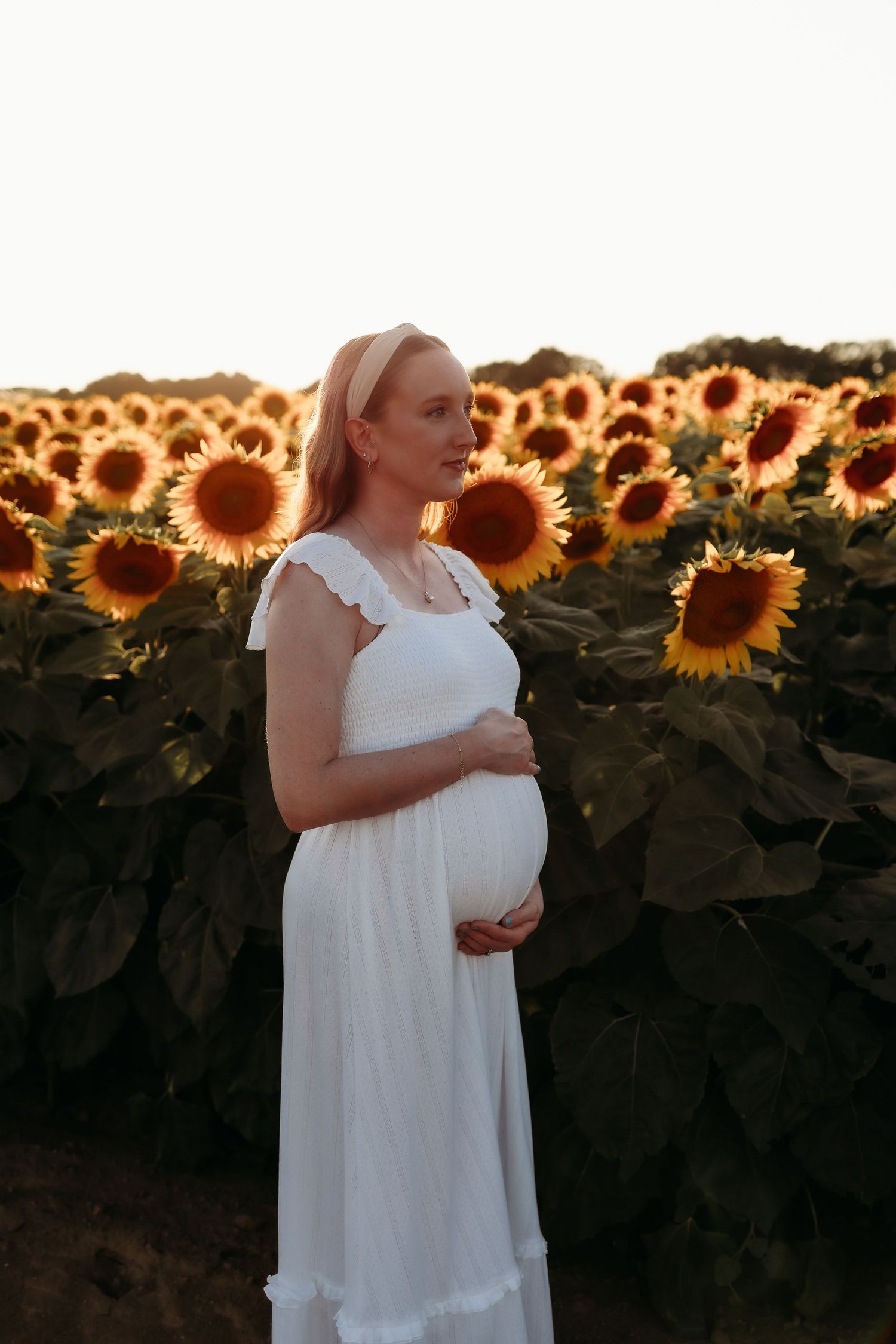 Pregnant blonde woman in white dress holds baby bump while standing in sunlit sunflower field at dusk.
