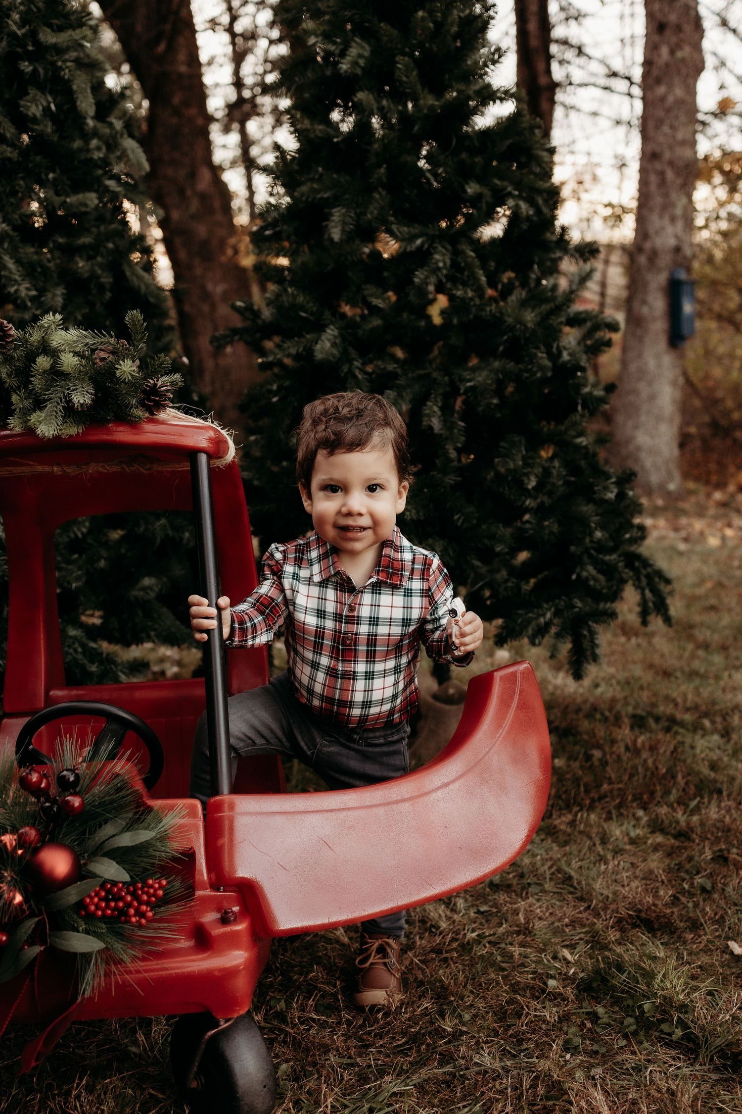 Young boy in plaid shirt sitting in red vintage toy truck near Christmas trees outdoors.