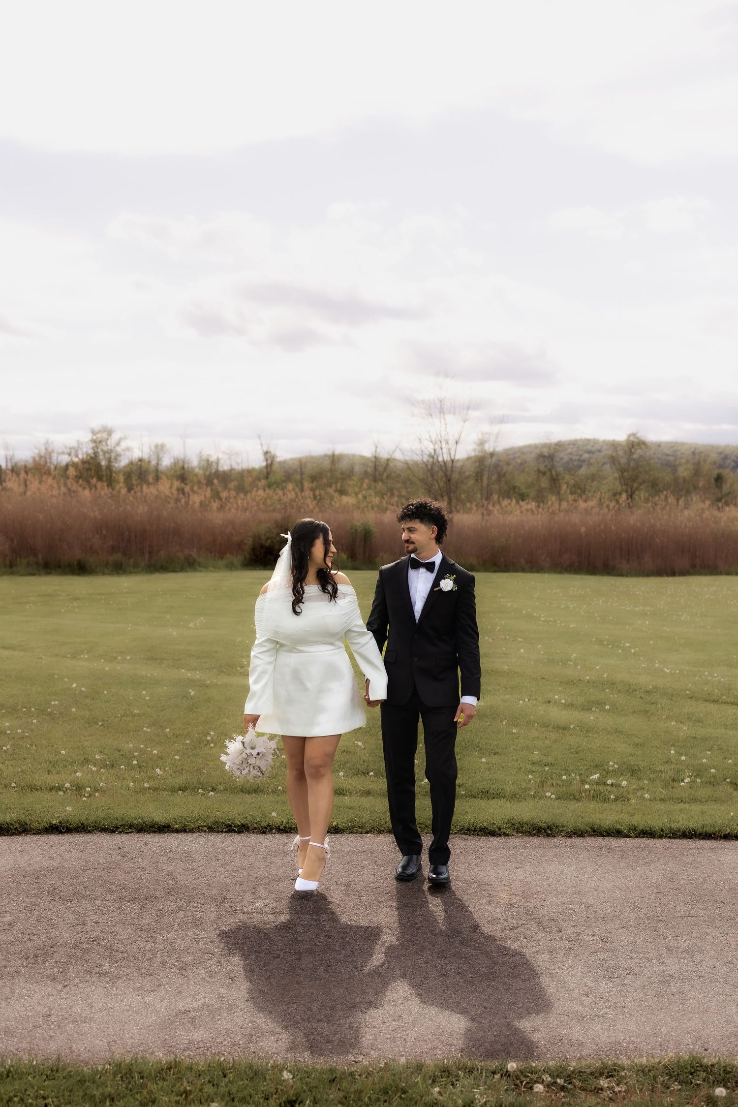 Bride in white mini dress and groom in black tuxedo walking hand in hand on a path with green fields behind.