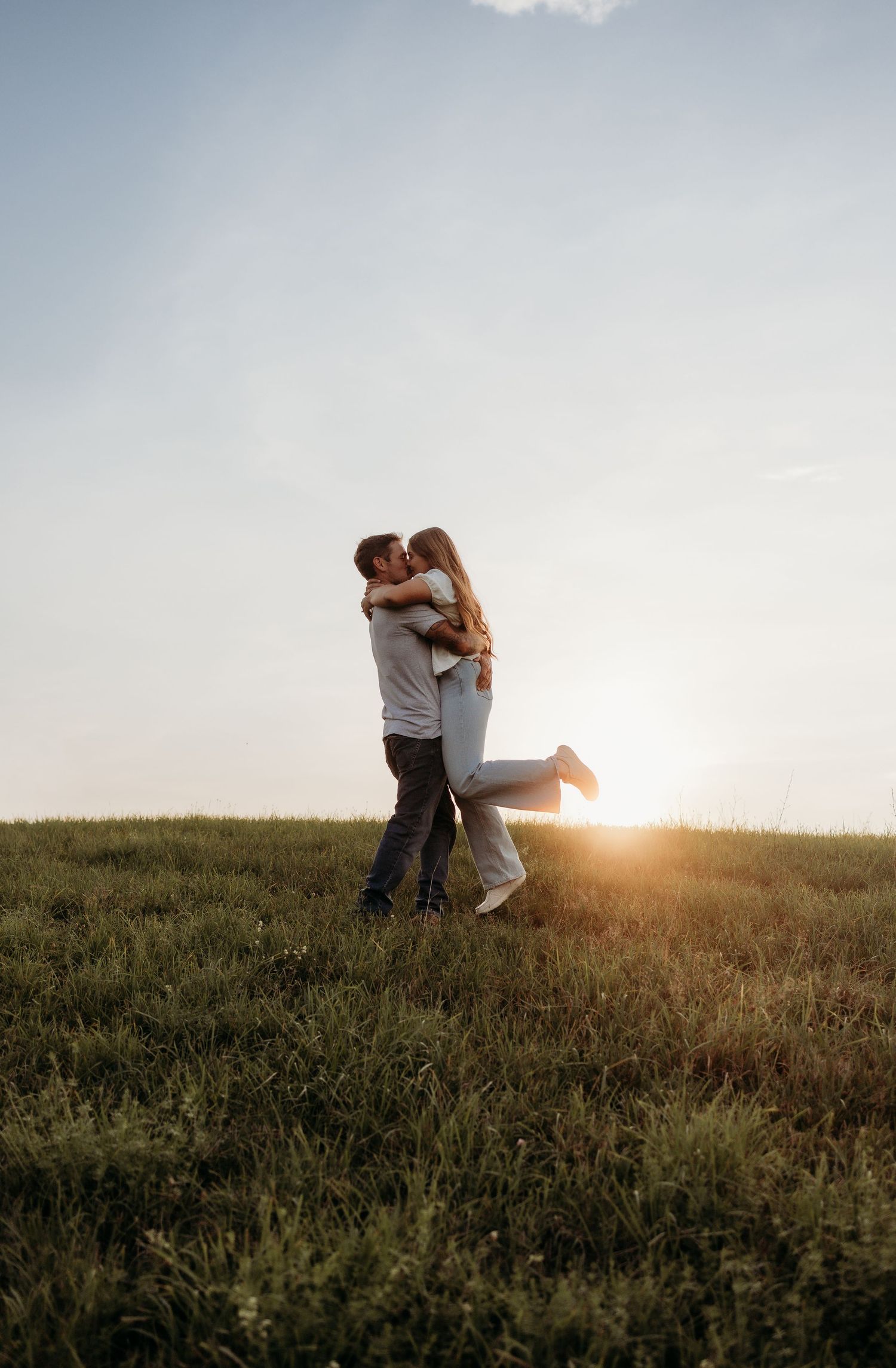 A couple shares a romantic embrace and kiss in a sunlit green meadow at golden hour.