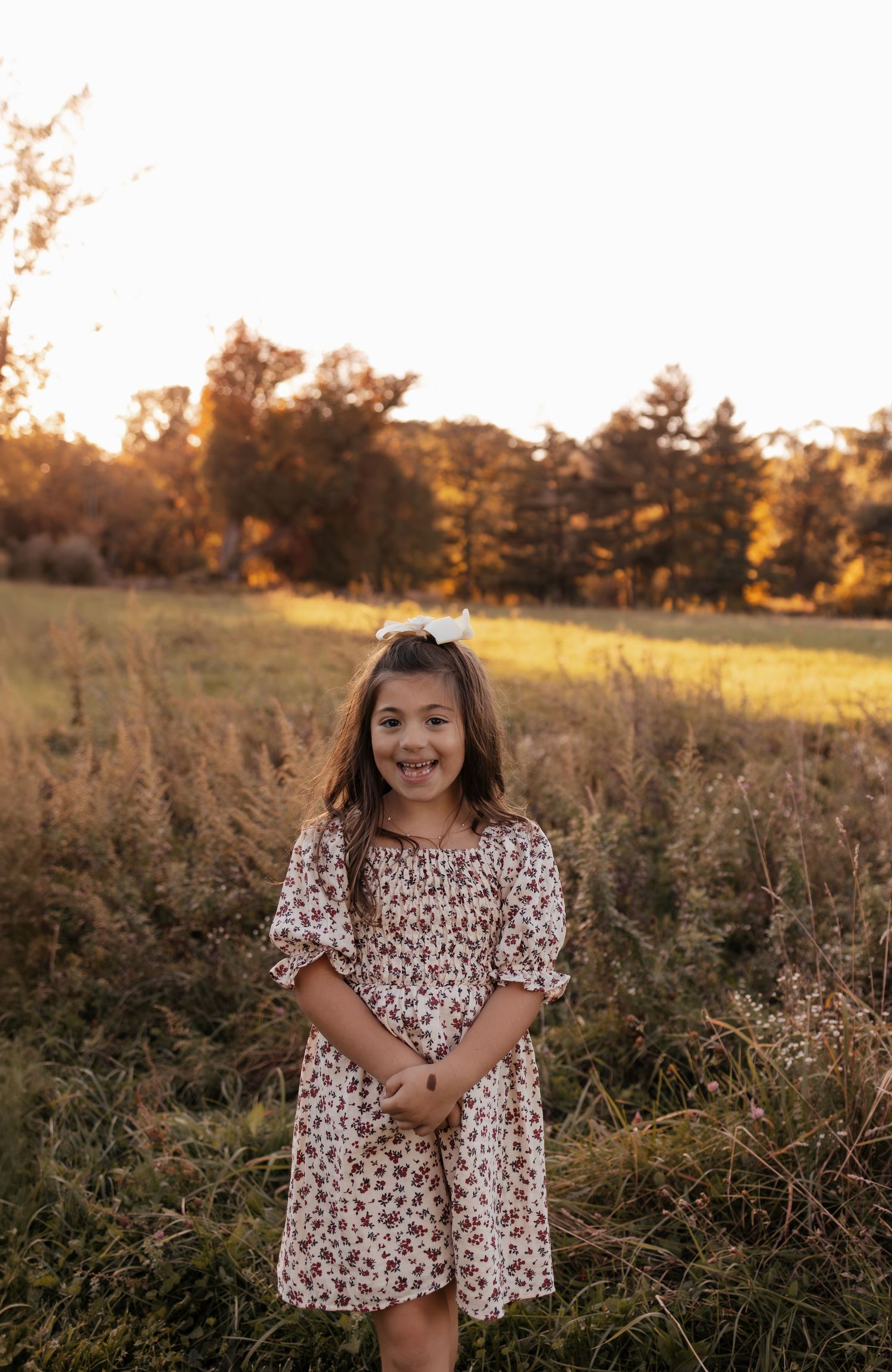 Smiling girl in a floral dress and white hair bow stands in a golden grassy field at sunset with trees in the background.