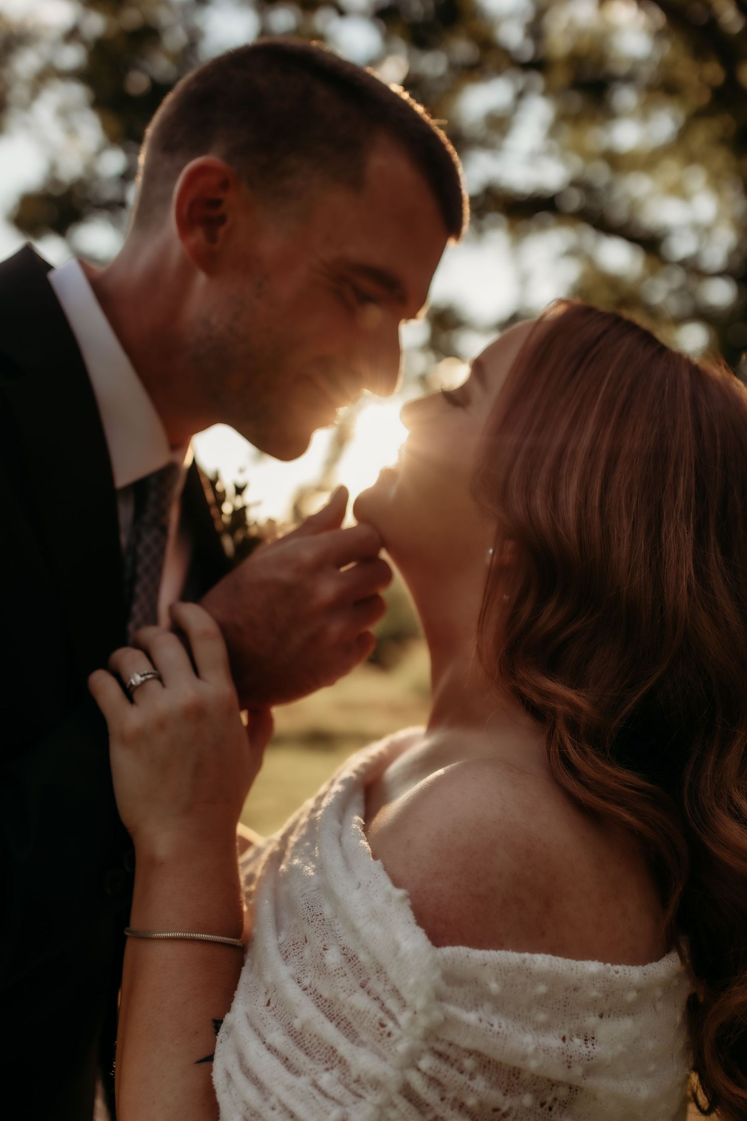 A bride and groom share a romantic golden hour moment, sun glowing between them as she touches his face in wedding attire.
