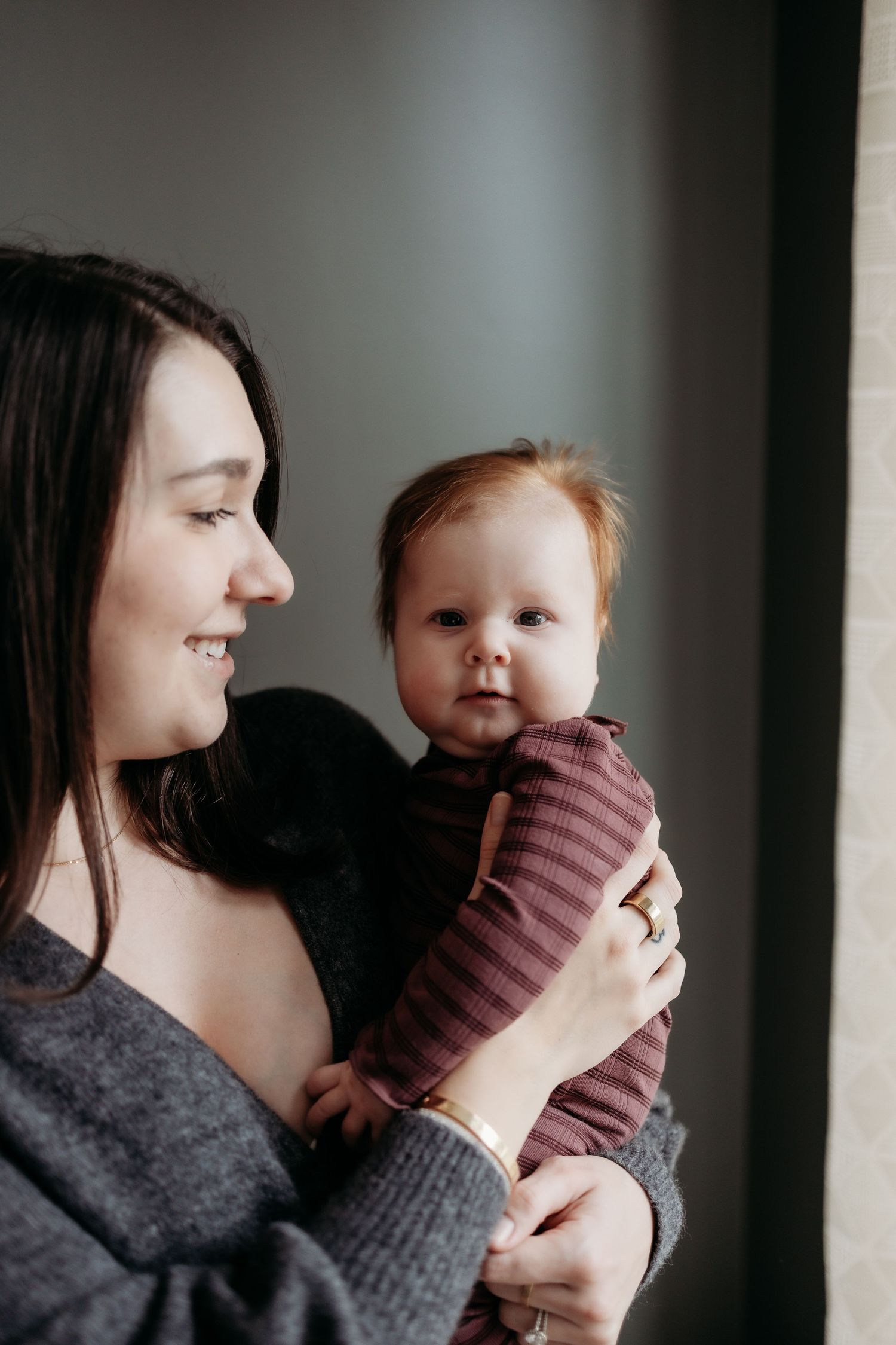 Smiling young mother holding her red-haired baby in a striped outfit near a window.
