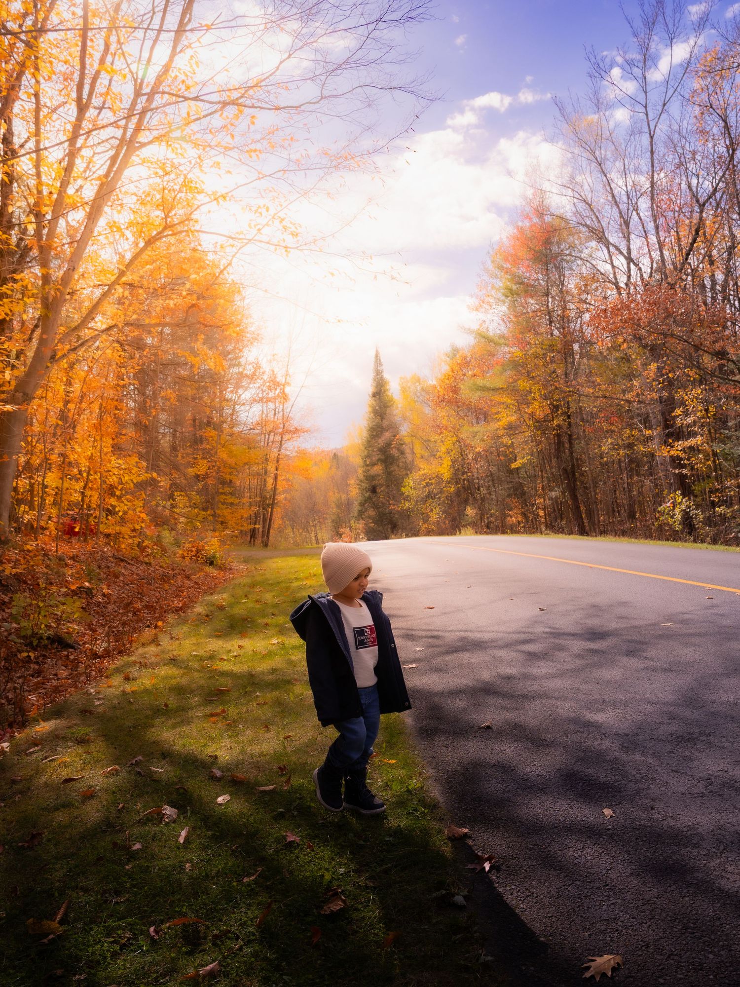 Young child in winter coat stands beside scenic autumn road with colorful fall foliage.