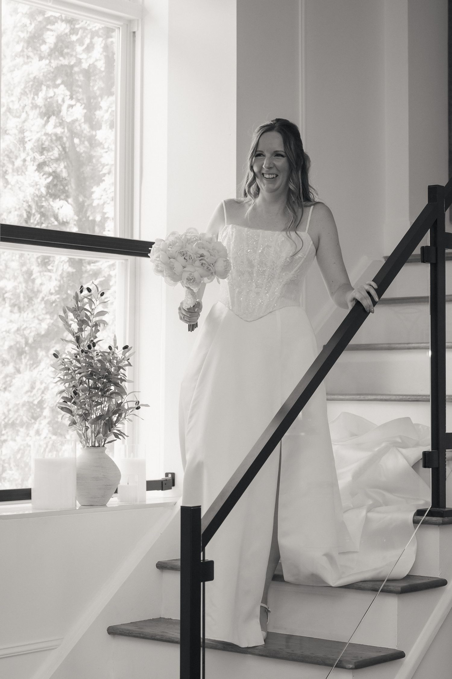 Black and white photo of smiling bride descending staircase in lace gown holding white floral bouquet.