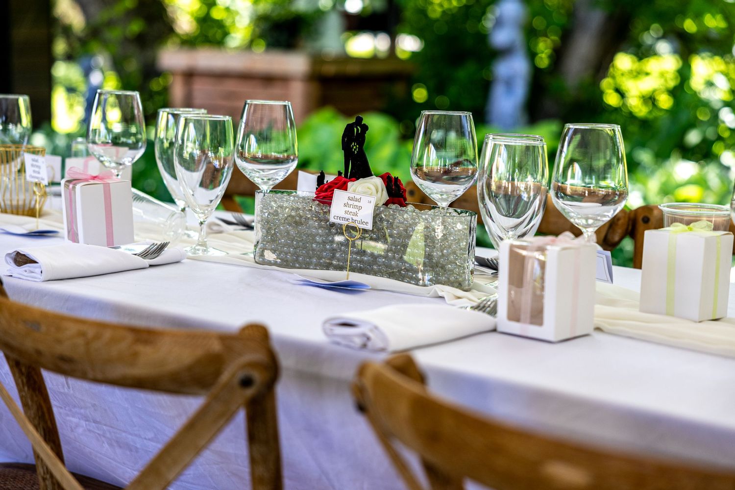 Elegant outdoor wedding reception table set with crystal wine glasses, white linens, and decorative favor boxes.