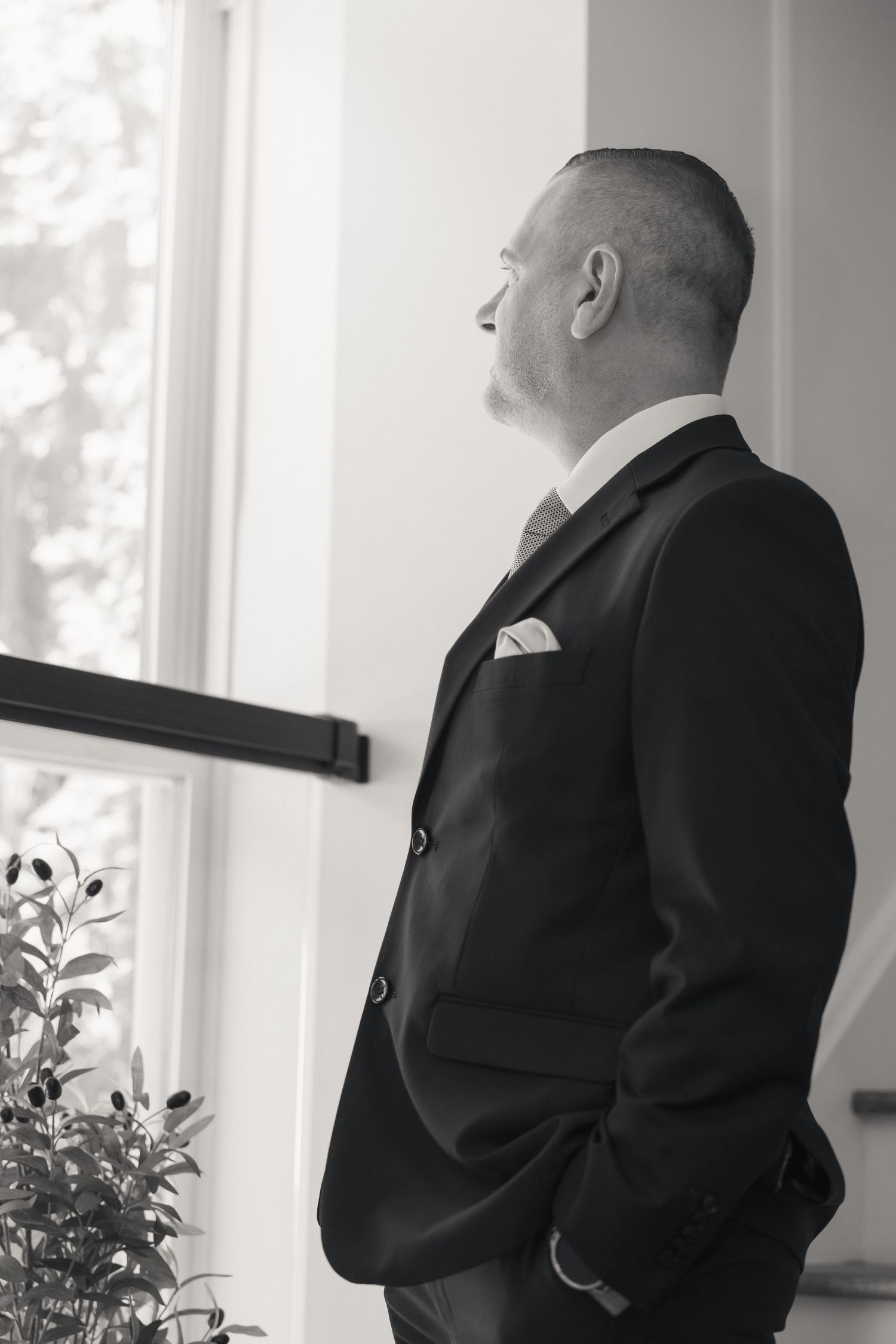 Black and white portrait of groom in navy suit gazing thoughtfully out a bright window.
