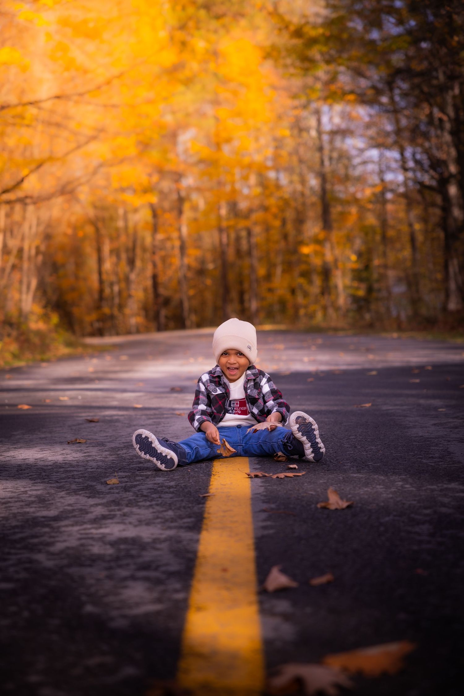 Happy child in plaid shirt and beanie sits laughing on road surrounded by autumn leaves.
