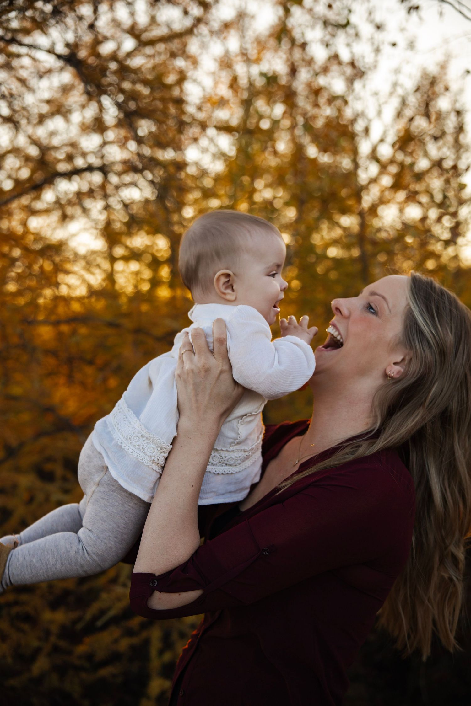 A joyful mother lifts her smiling baby outdoors in a golden autumn forest setting.