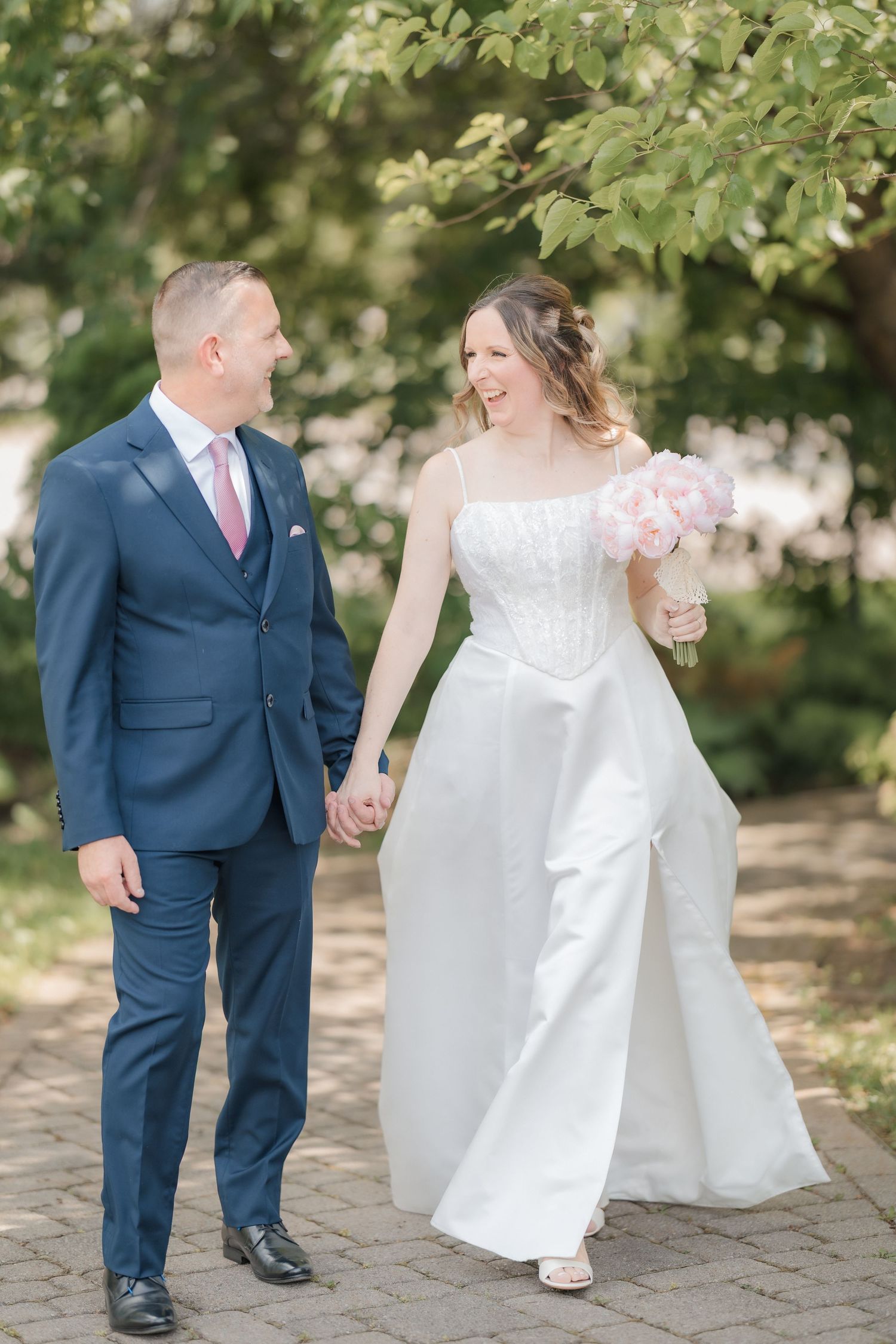 Bride and groom walking hand in hand through tree-lined path, bride carrying pink peony bouquet.
