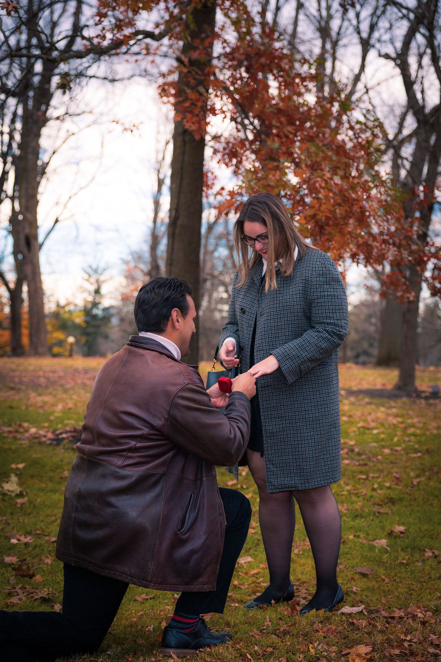 Man kneels to propose to woman in gray coat amid colorful autumn park trees.