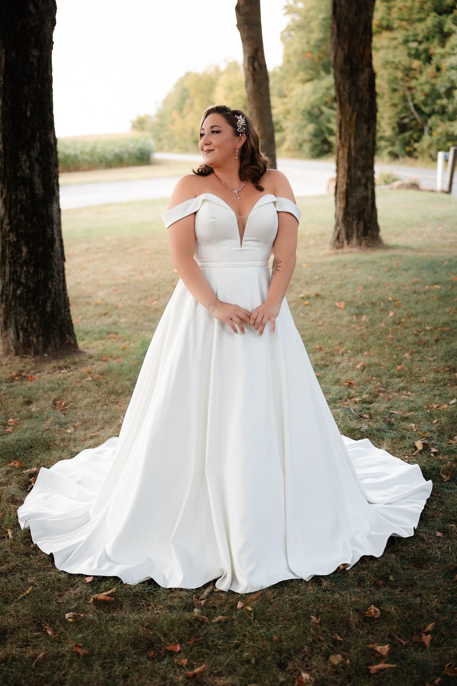 Plus-size bride in off-shoulder white satin ball gown with deep V-neckline posing between trees outdoors.