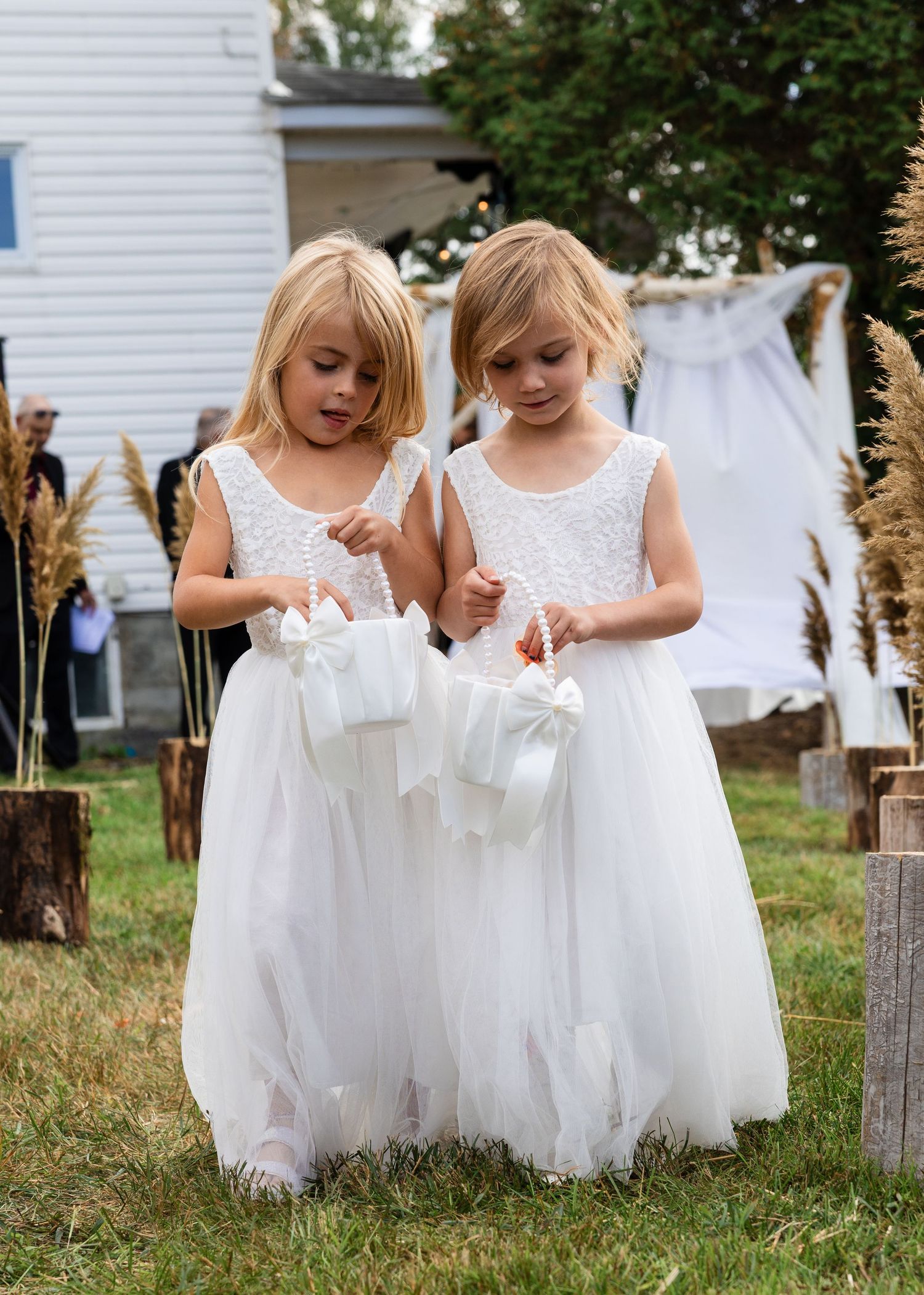 Two adorable flower girls in white tulle dresses carrying flower baskets at outdoor wedding ceremony.