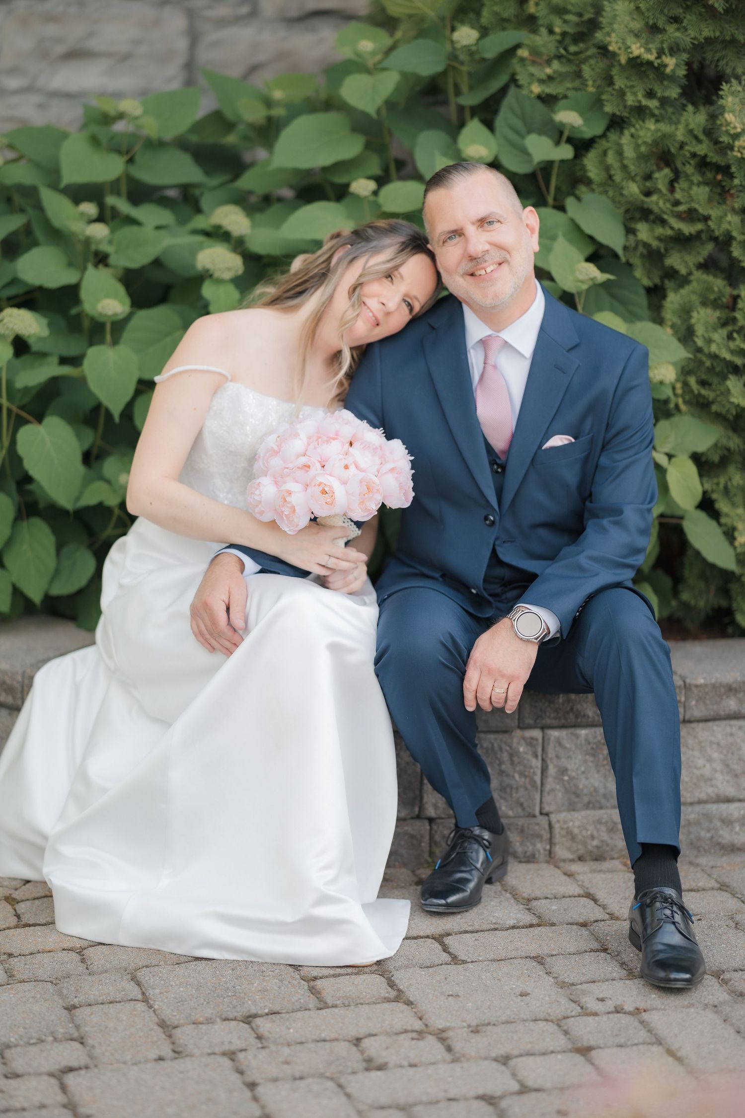 Bride and groom sitting together on stone steps, bride holding pink peony bouquet, groom in navy suit with pink tie.