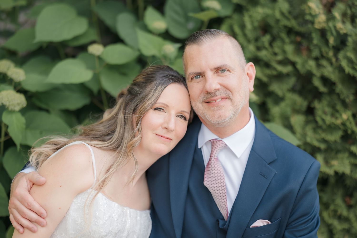 Bride and groom posing together smiling in front of lush green foliage.